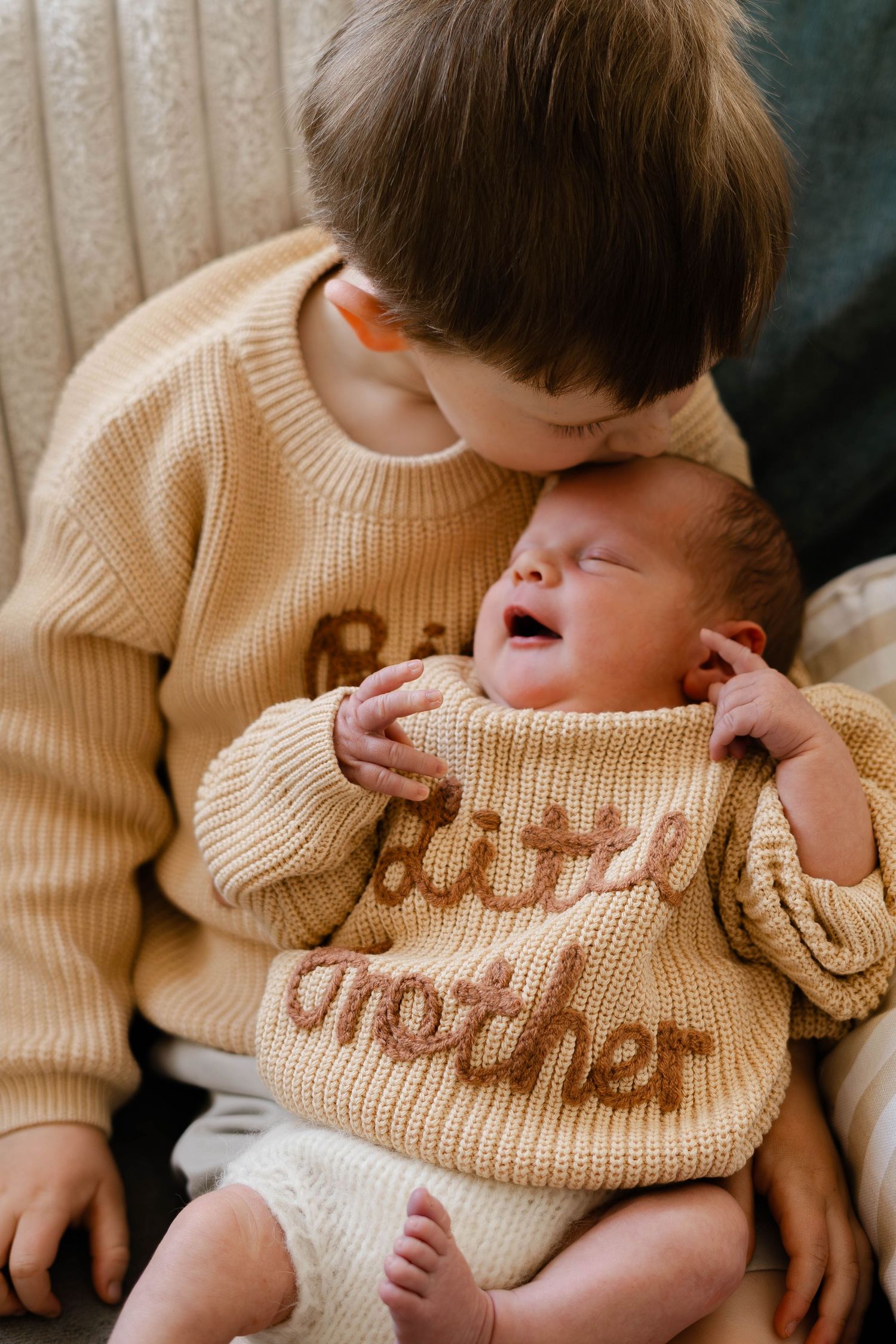 Sibling cuddles newborn wearing cream knit sweater during lifestyle photo session.