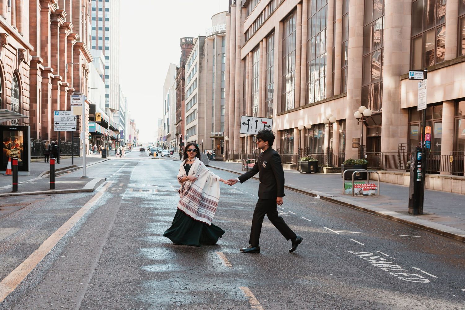 Couple crossing an empty urban street, woman in flowing white dress, man in dark suit, holding hands in city.