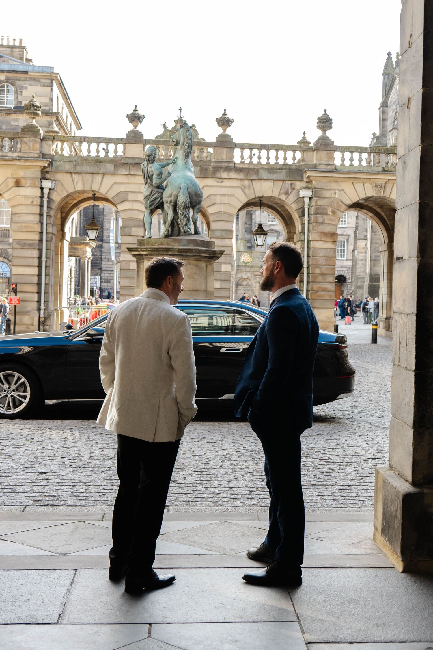 Two people in formal attire stand in conversation near a historic building with ornate architecture.