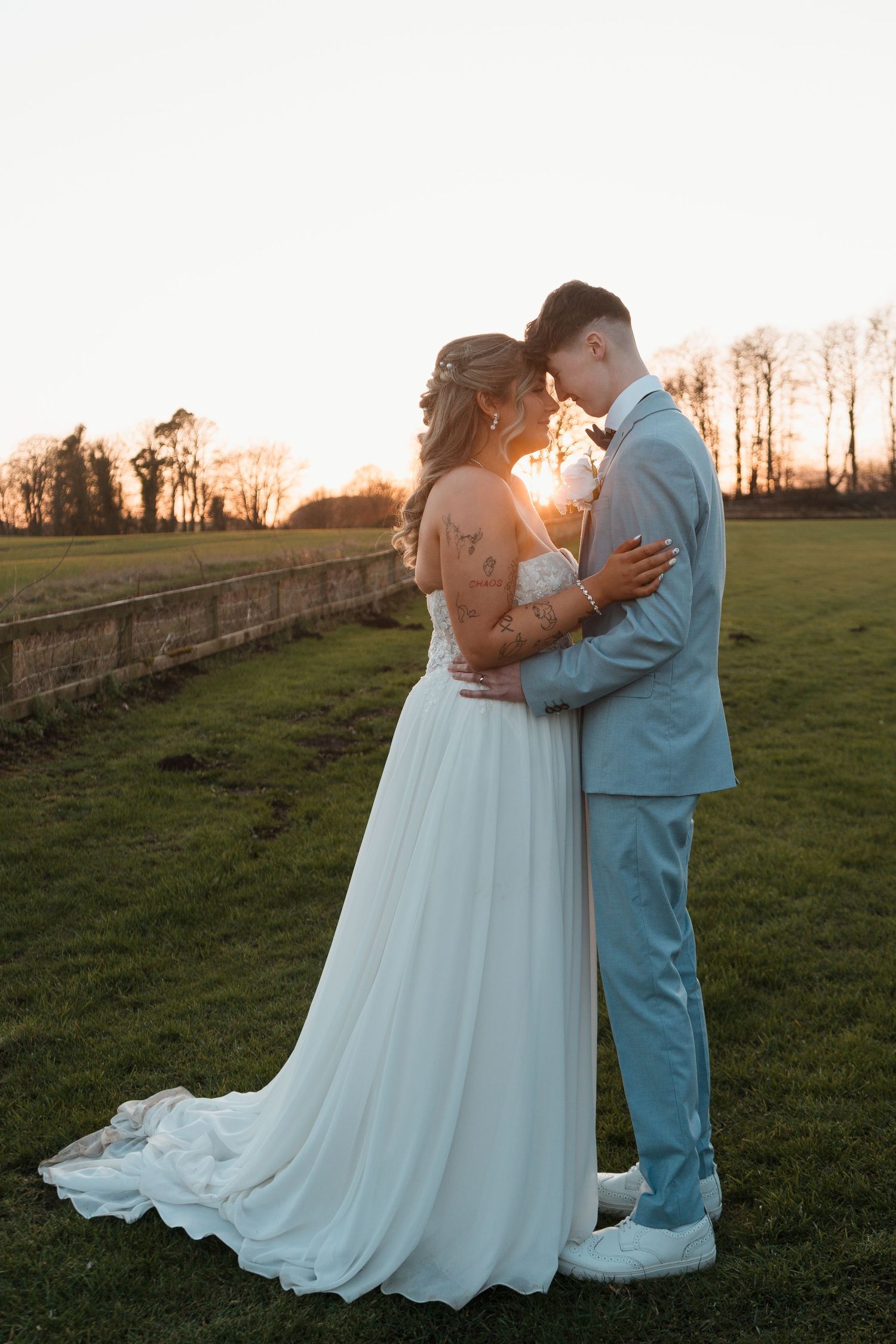 Bride and groom embrace at golden hour sunset on green lawn, she in white gown, he in light blue suit.
