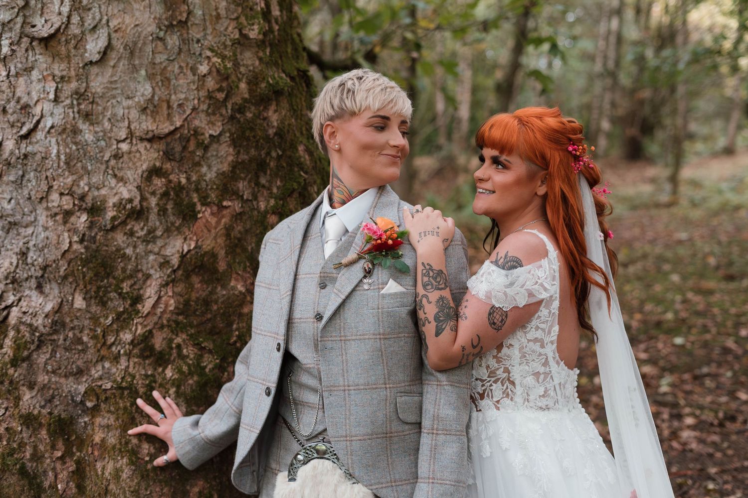 Bride and groom pose by a tree in a forest, she wears lace gown with veil, he wears grey plaid suit.