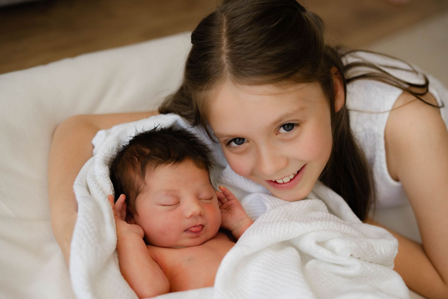 A child smiles while cuddling with a sleeping newborn on white bedding.