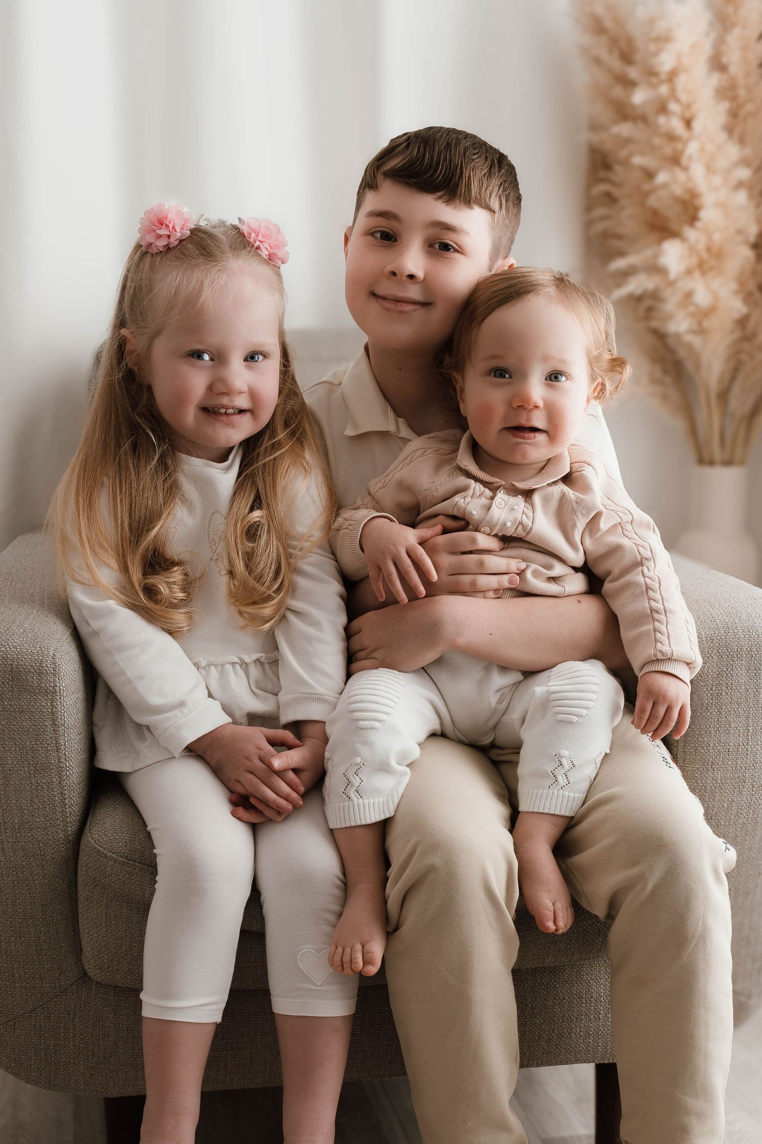 Three young siblings pose together on a gray chair, dressed in neutral tones, smiling warmly in a cozy indoor setting.