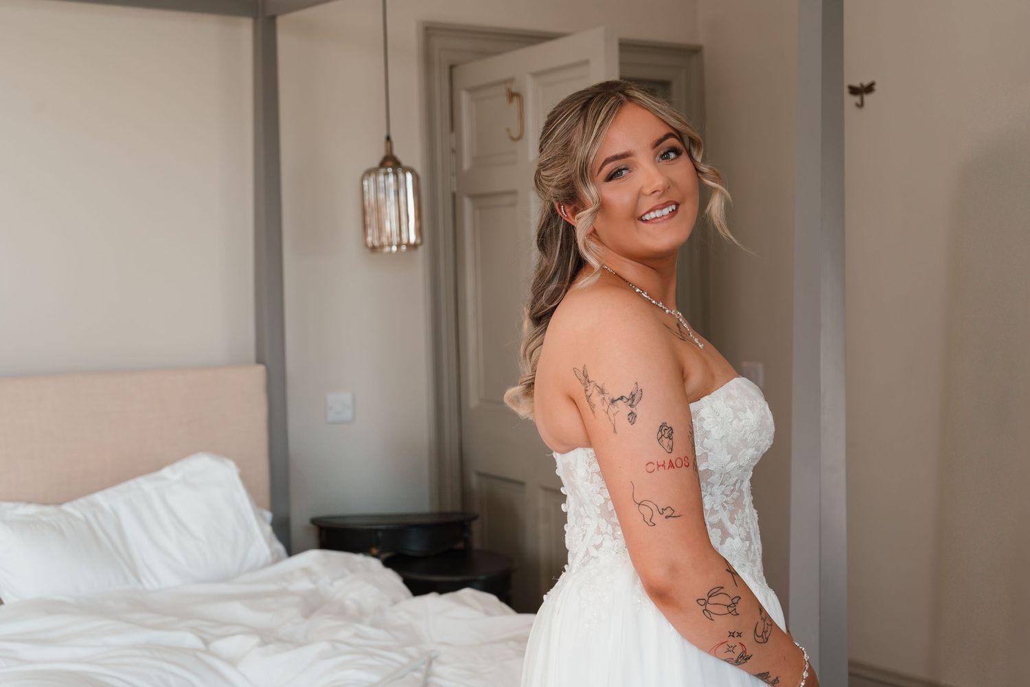 Smiling bride in a strapless lace wedding gown with tattoos, posing in a bright bedroom.