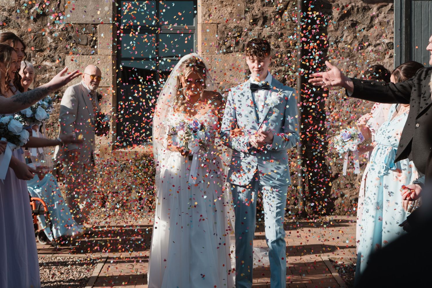 Bride and groom walk through colorful confetti shower surrounded by guests at outdoor wedding ceremony.