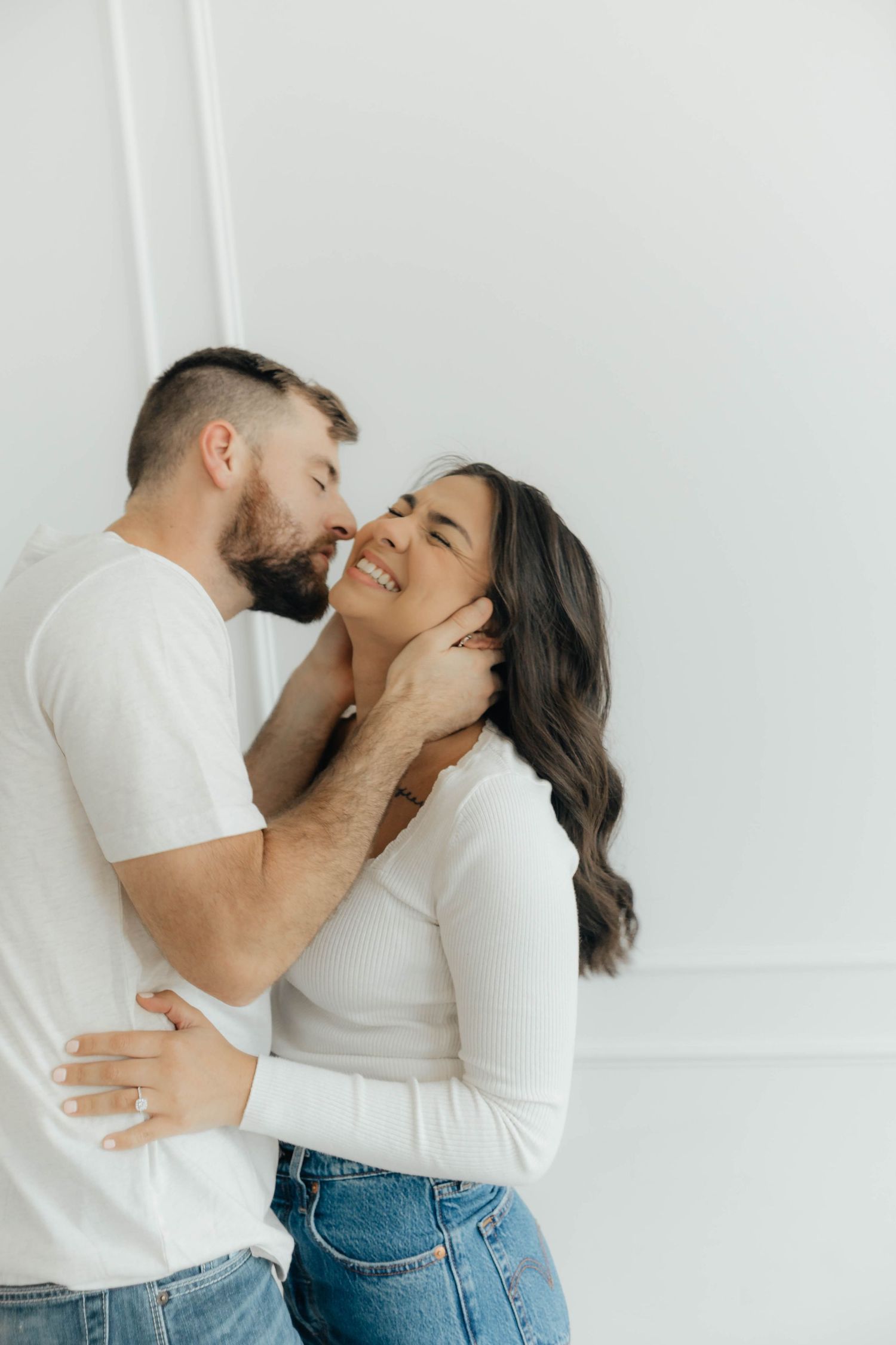Intimate couple sharing a tender moment against a white wall in casual attire.
