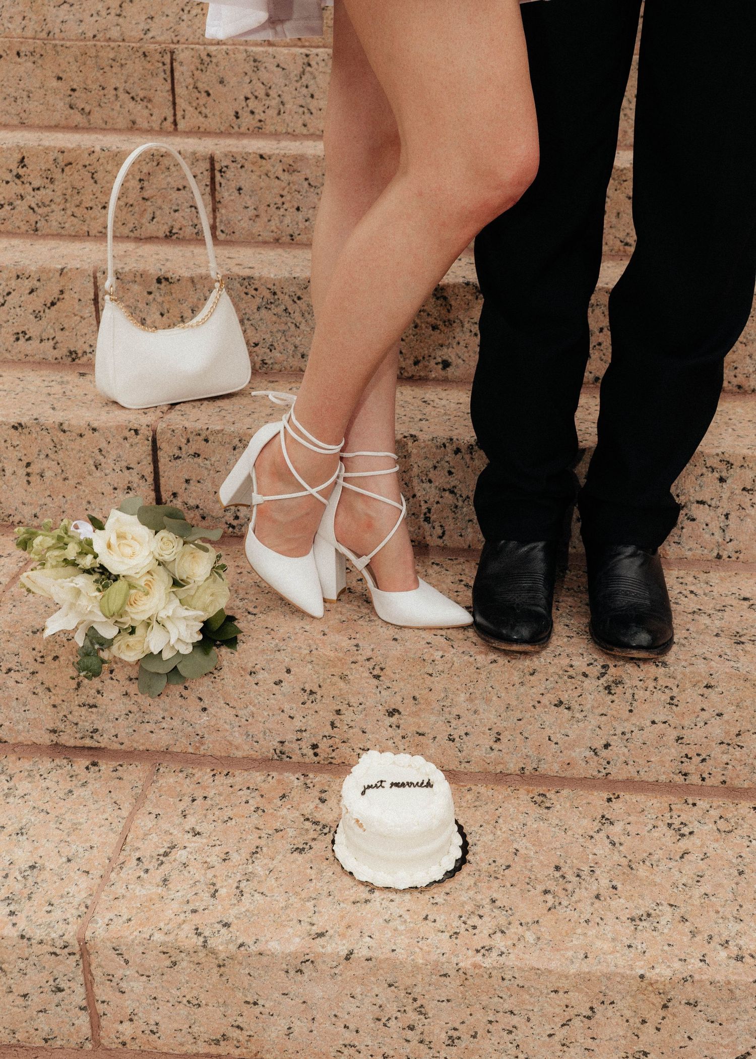 Bride and groom standing on stone steps with small wedding cake, white heels, floral bouquet, and white handbag.
