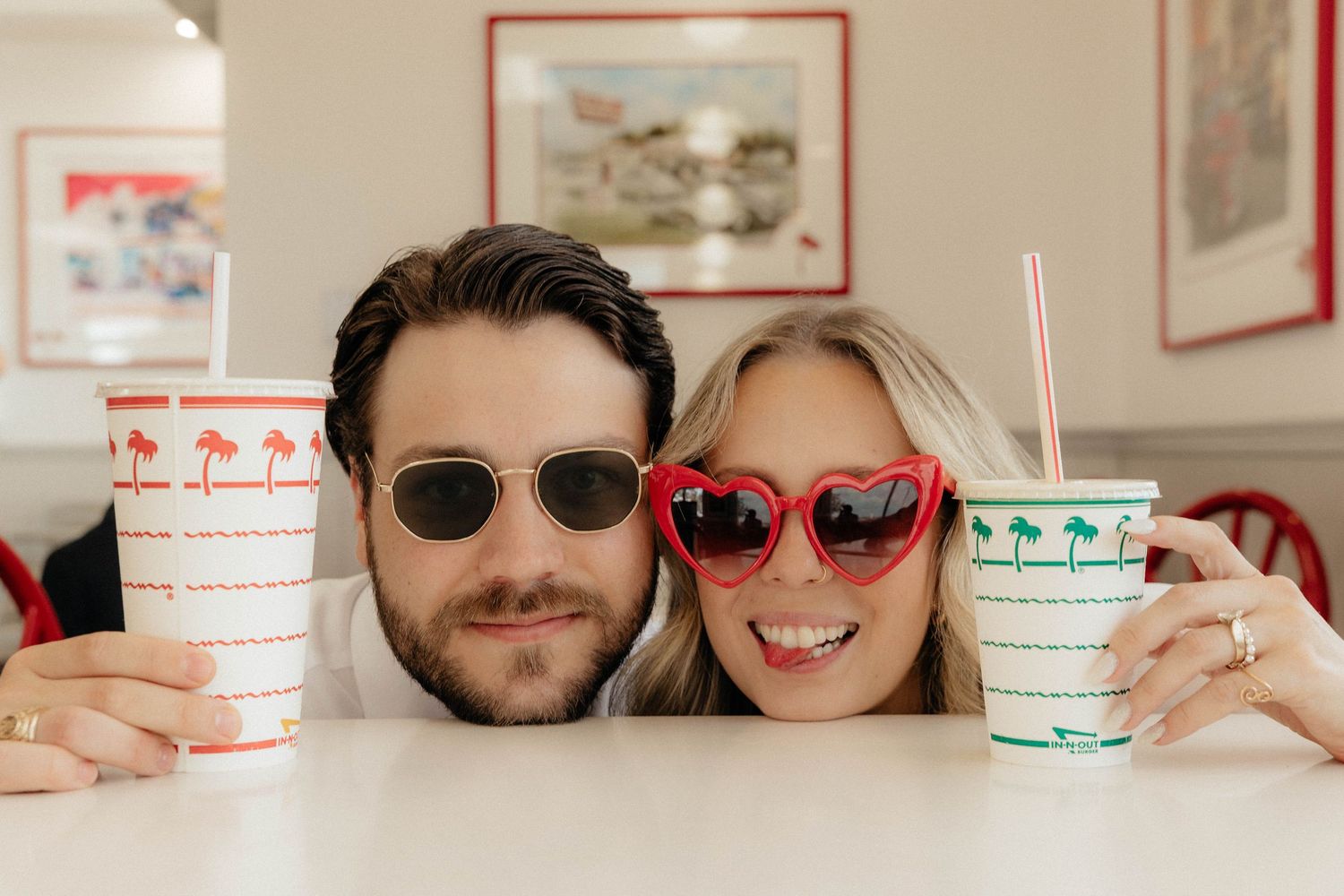 Couple smiling at camera holding drinks in red and white cups, wearing sunglasses at a retro diner.