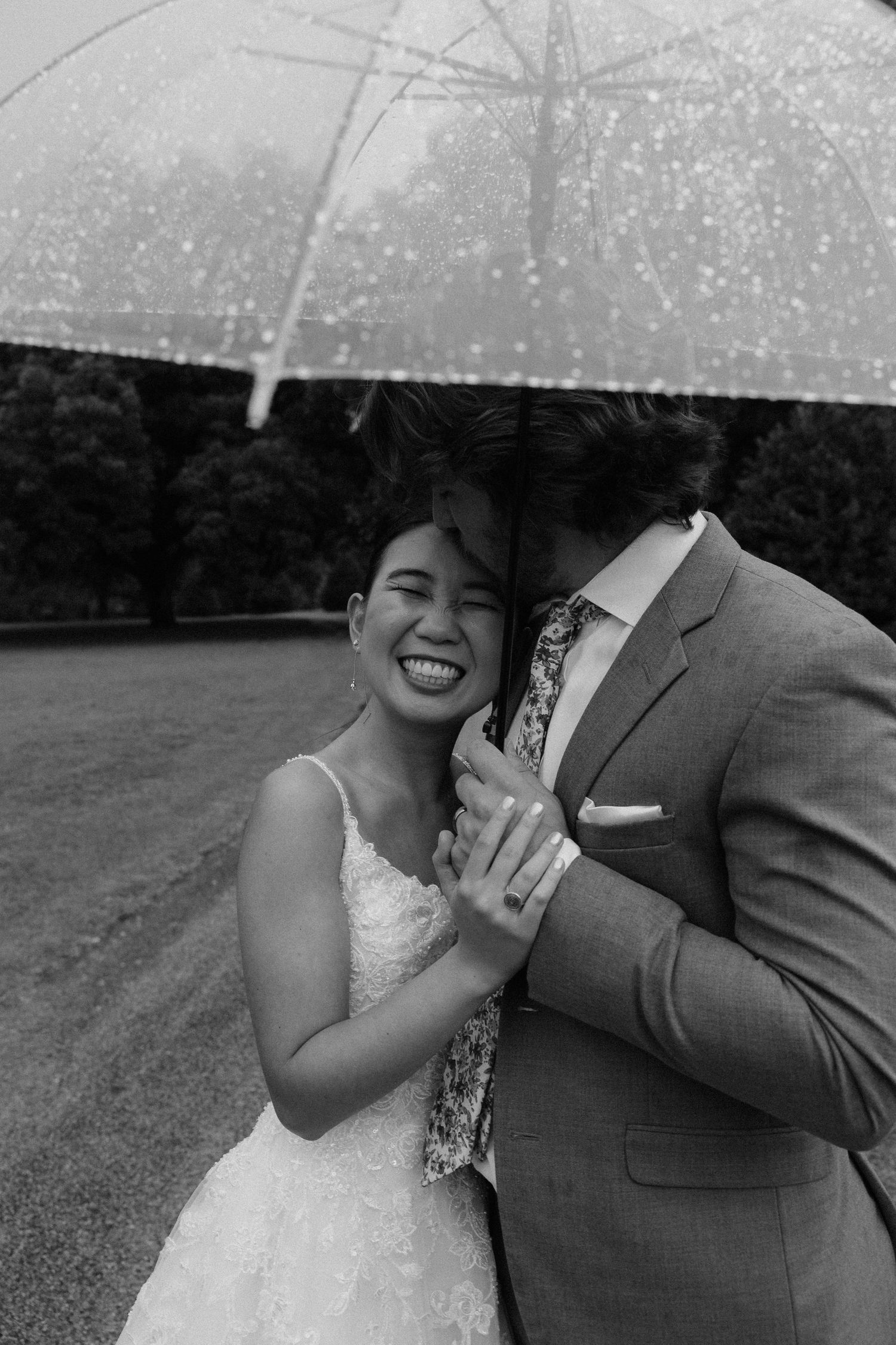 Black and white photo of a smiling bride and groom sharing an umbrella in the rain on their wedding day.