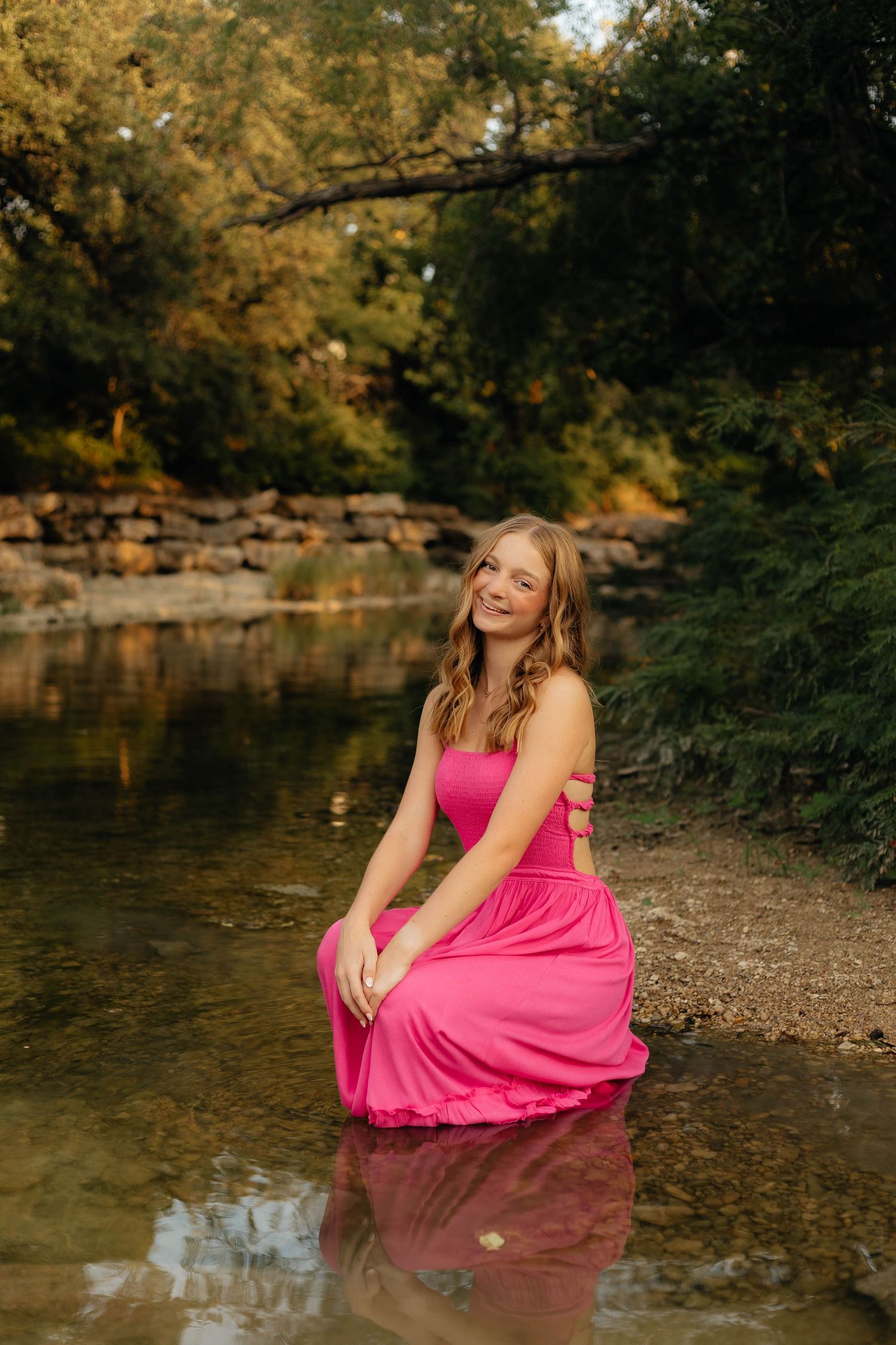 Woman in bright pink dress seated by reflective pond during autumn sunset.