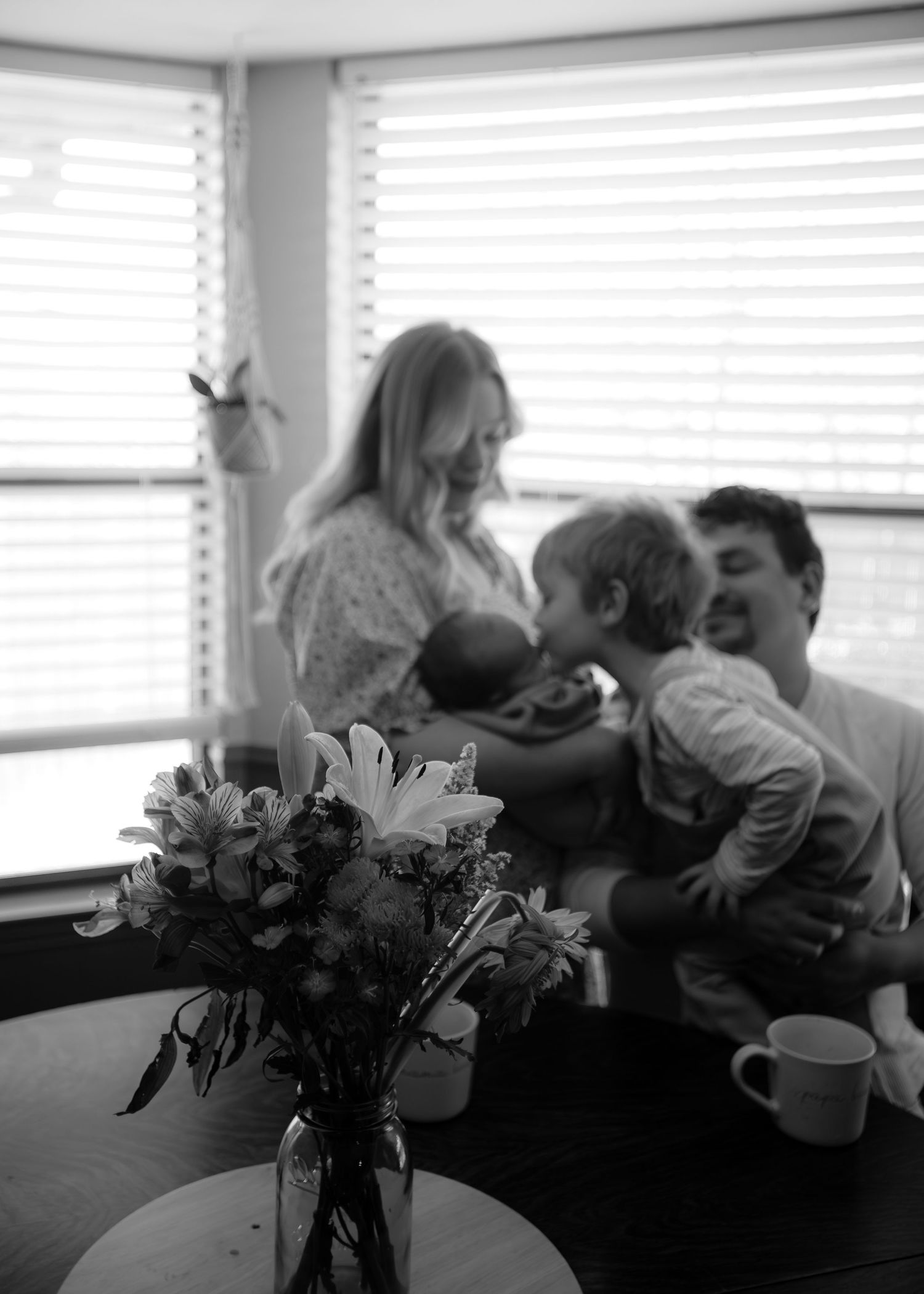 Black and white family photo with newborn baby, parents and toddler sharing a tender moment, flowers in foreground.