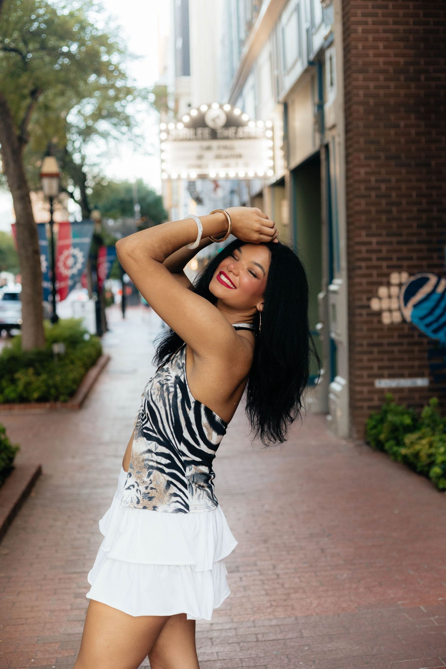 Woman in black and white outfit dancing joyfully on brick sidewalk near storefronts.
