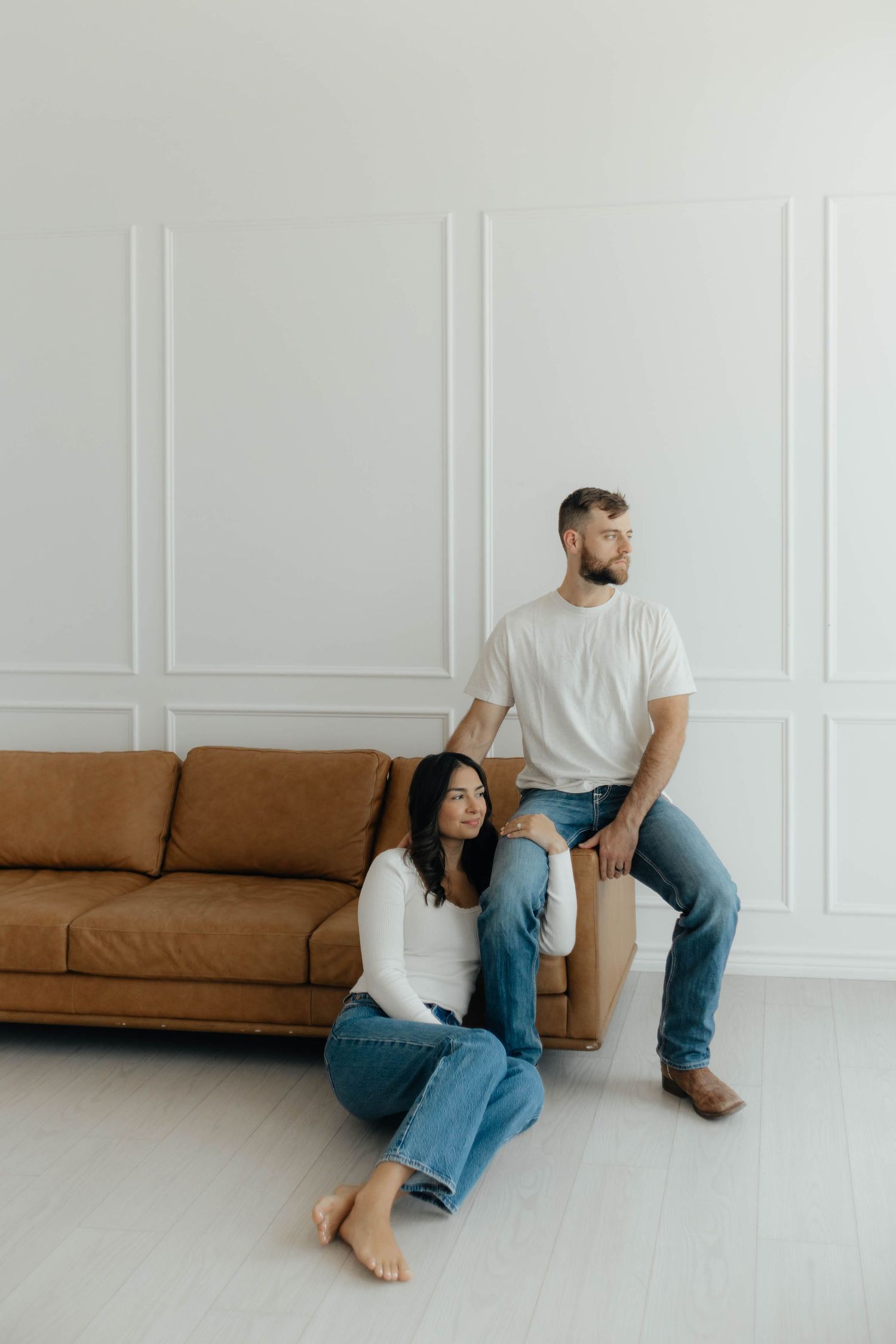 Couple relaxes casually on a brown leather couch in a minimalist white room.