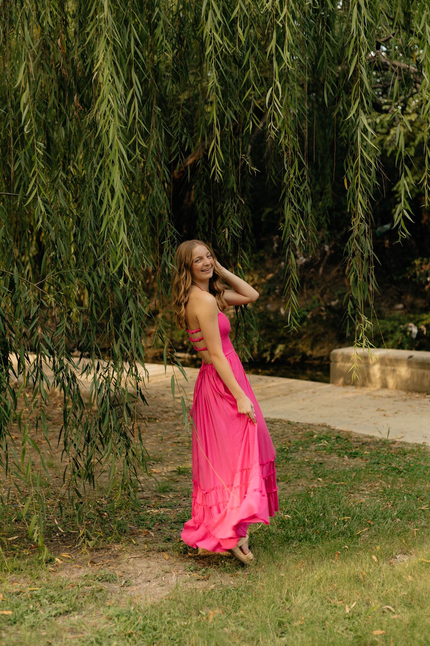 Young woman in a bright pink maxi dress smiling under a weeping willow tree in a lush green park.