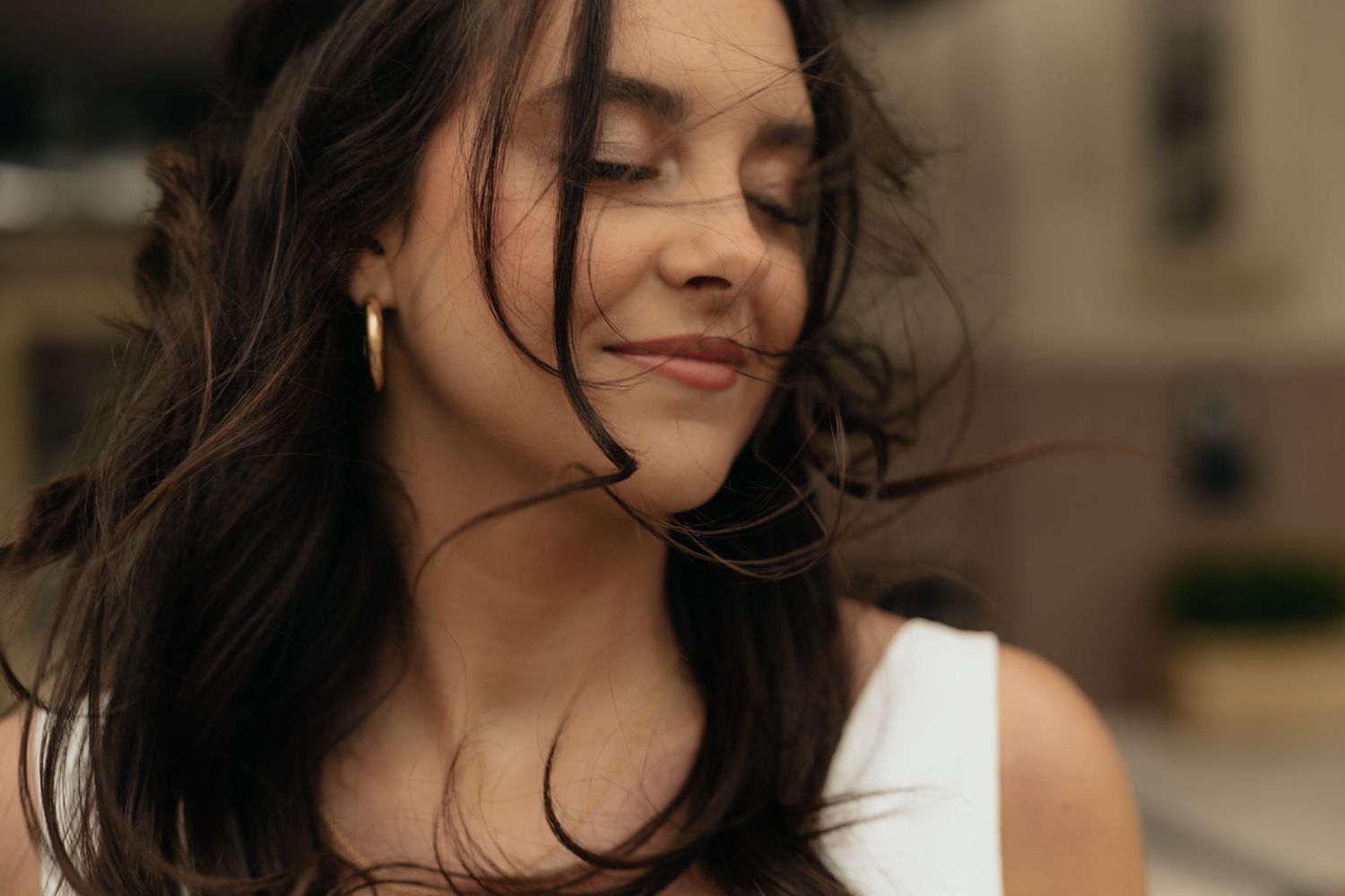 Young woman with dark hair smiling softly, eyes closed, wearing gold hoop earrings and white top outdoors.