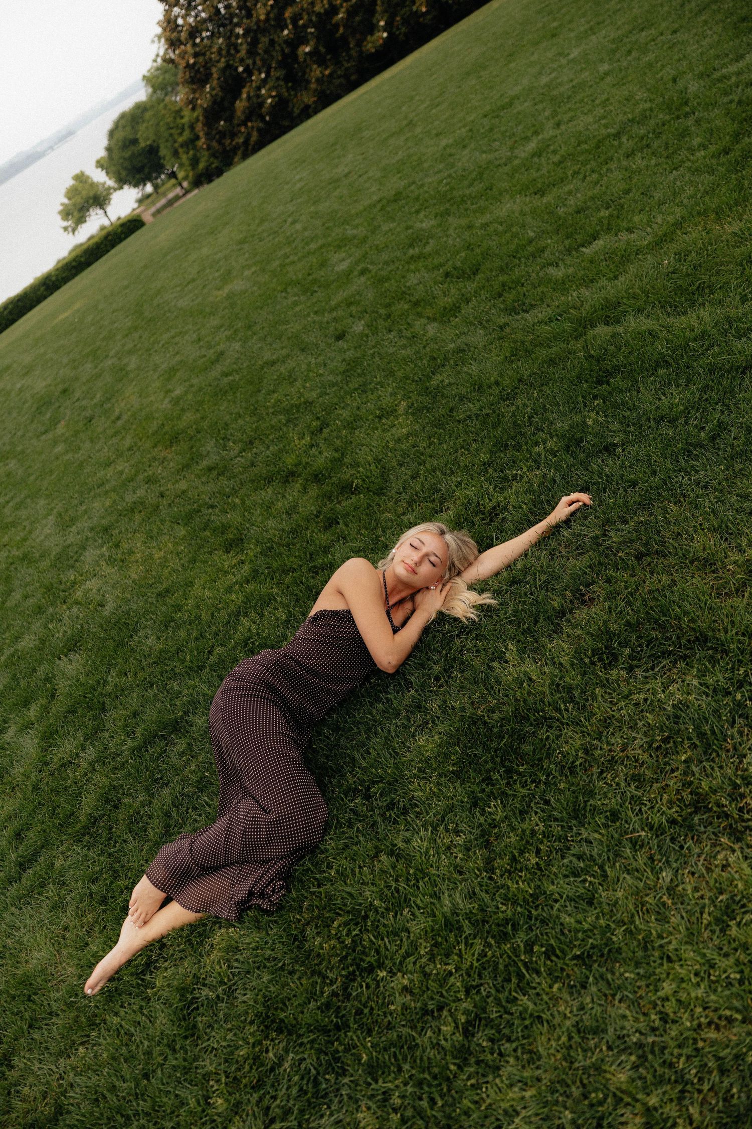A barefoot blonde woman in a dark dress lying on lush green grass, stretching her arm out in a serene outdoor setting.