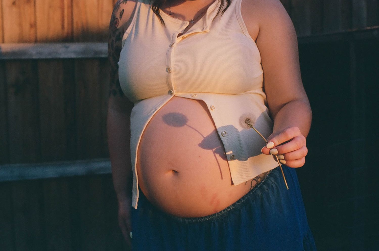 Pregnant woman in crop top and blue skirt showing baby bump outdoors in warm sunlight.