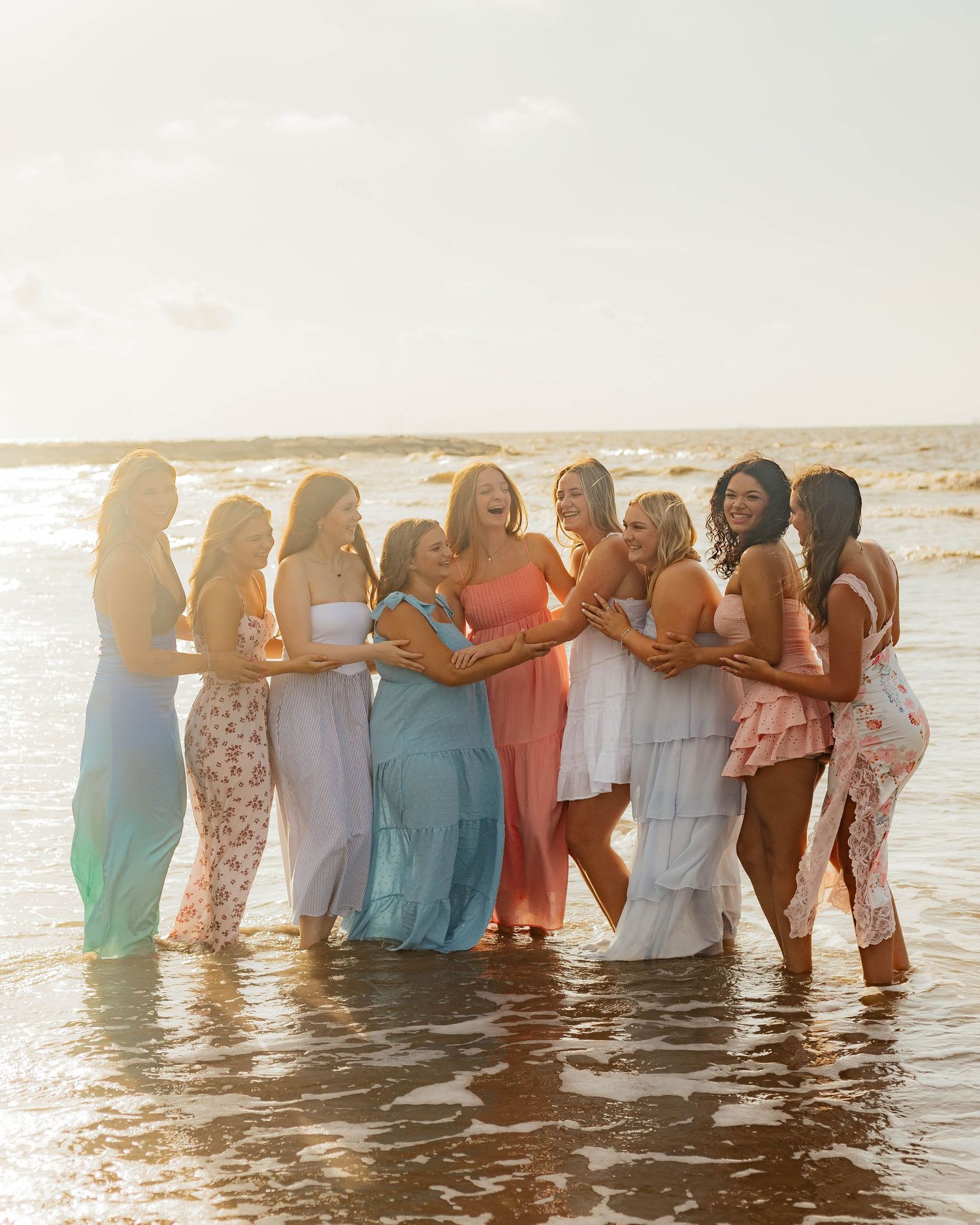 Group of friends in colorful dresses standing together in ocean water at sunset.