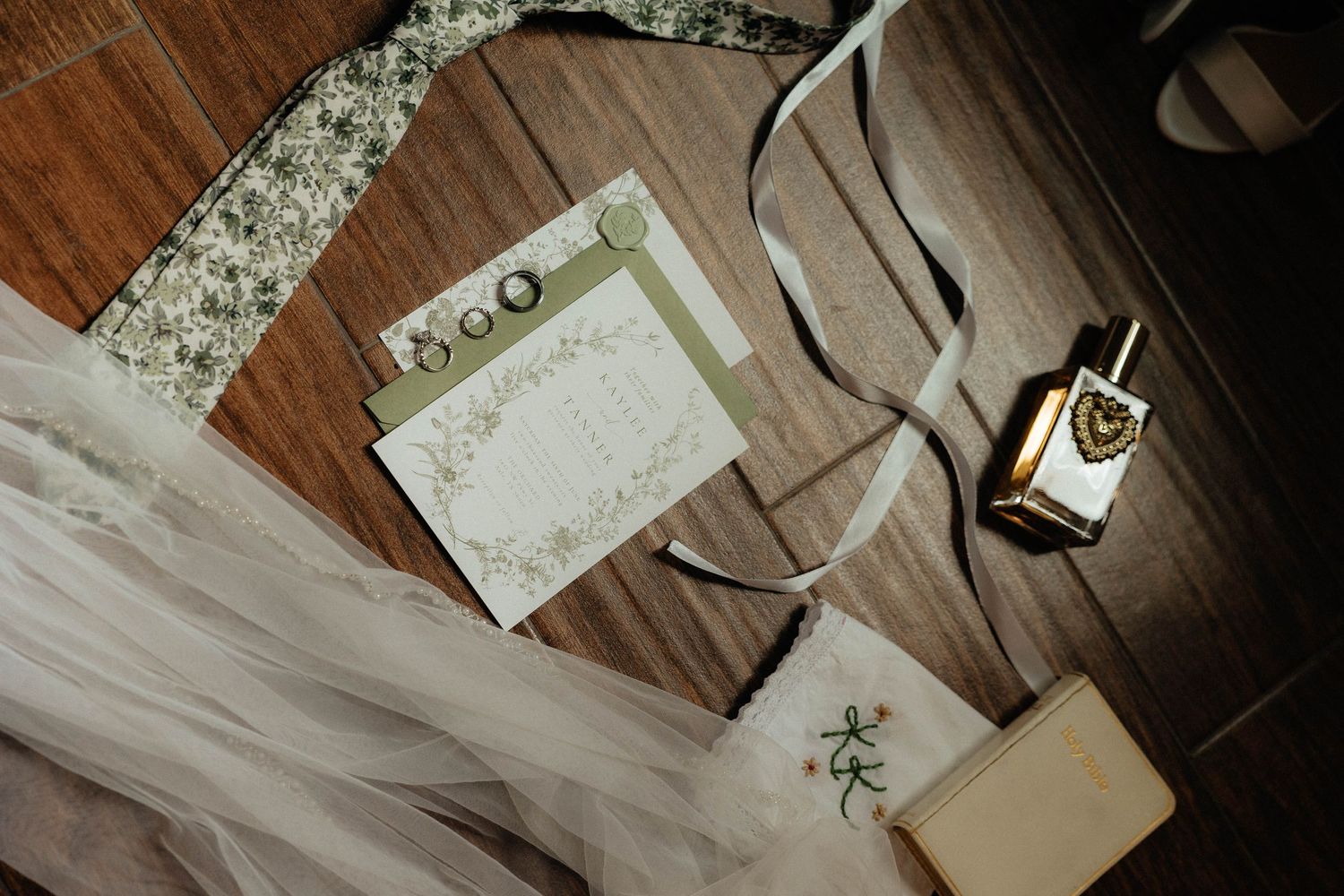 Elegant wedding flat lay with floral invitation, ribbon, perfume bottle, and bridal veil on wooden floor.