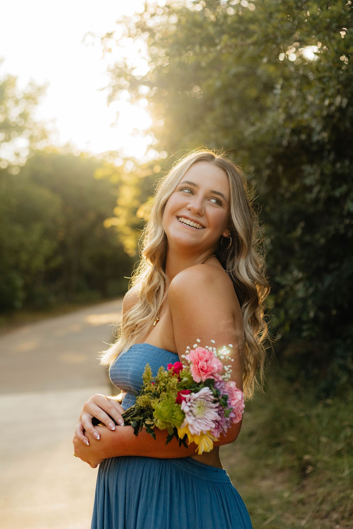 Woman in blue dress holding colorful flowers poses outdoors during golden sunset light.