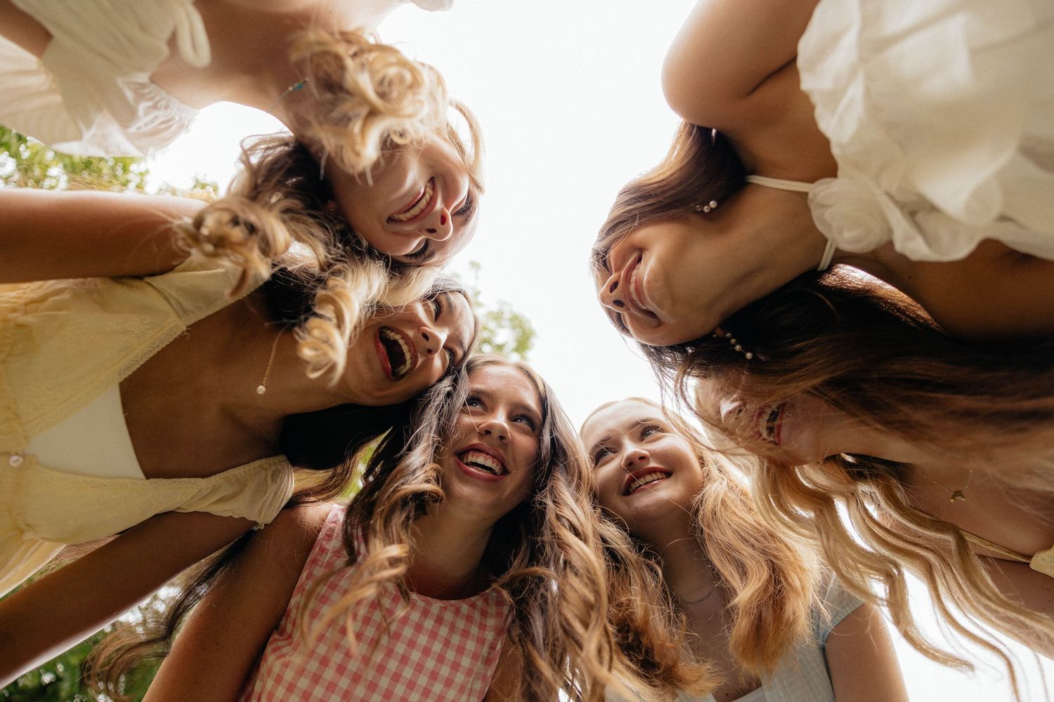 Six smiling young women huddle together in a circle, looking down at the camera on a sunny day.