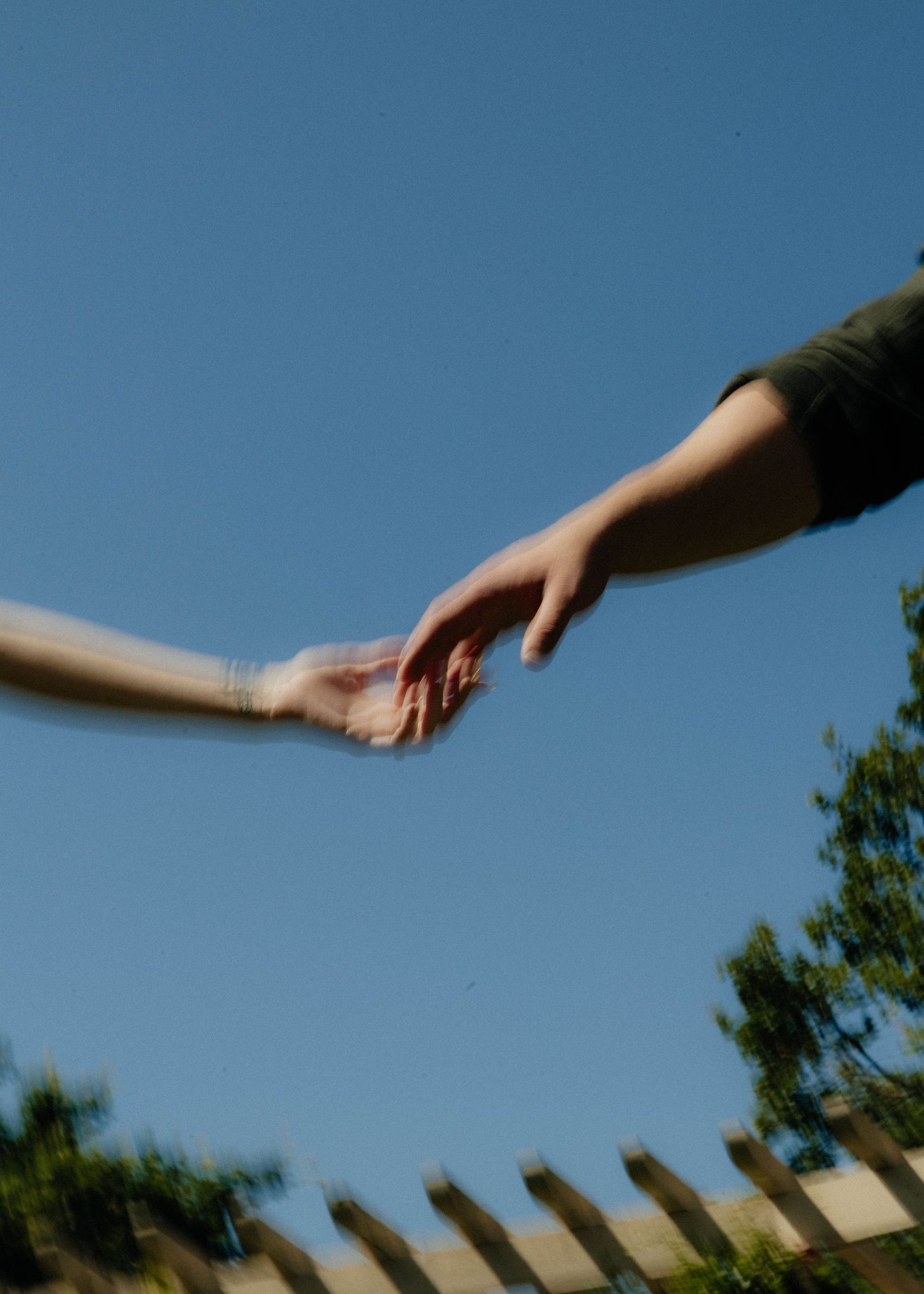 Two hands reaching toward each other against a clear blue sky with trees and a wooden fence below.