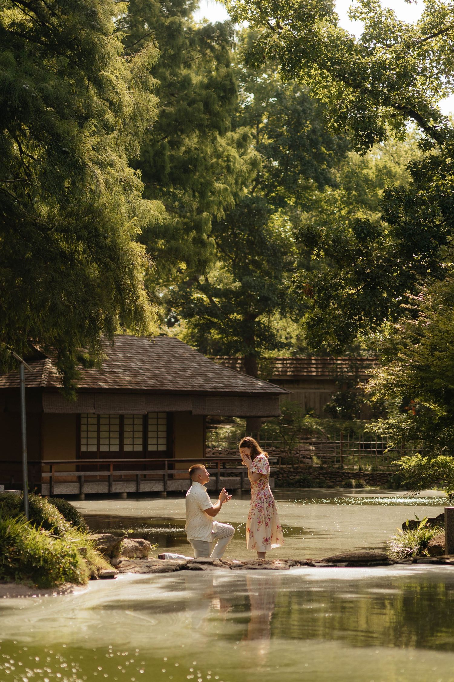 Man proposing to woman by a serene pond near a Japanese garden pavilion surrounded by lush green trees.
