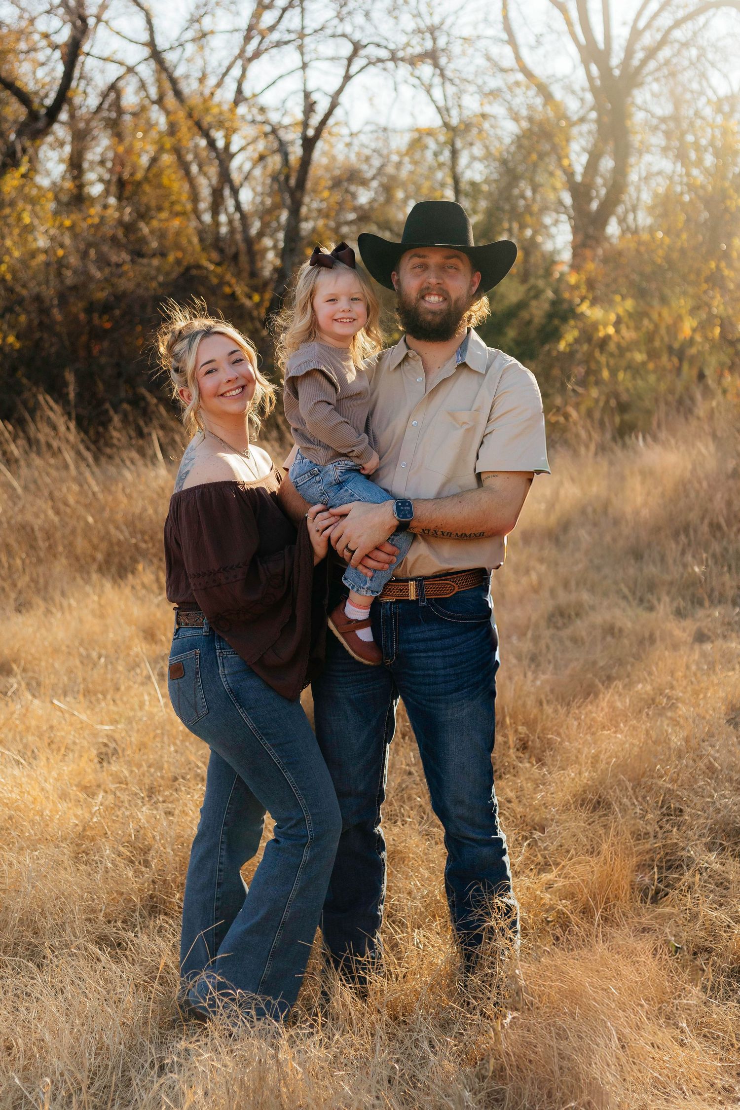 Family poses together in golden autumn field wearing western style clothing during sunset photo session.