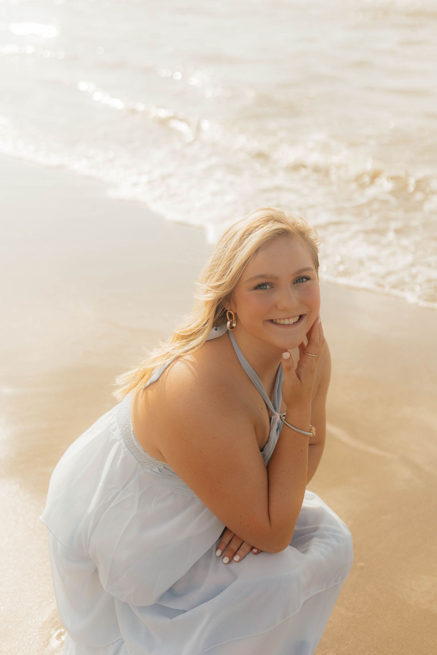 Woman in white dress sitting on beach sand during sunset with waves in background.