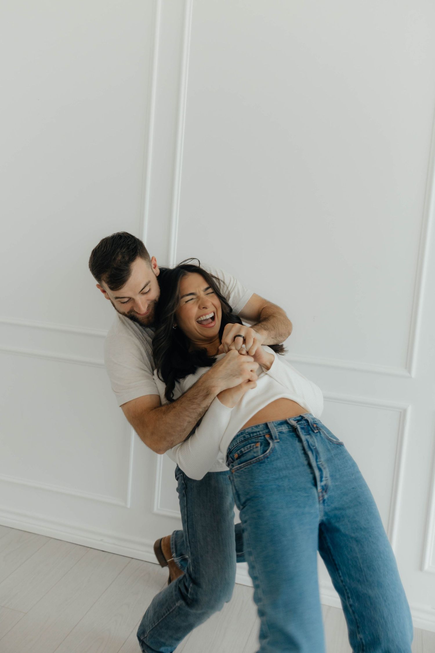 Couple in denim and white sharing a joyful embrace against white wall.