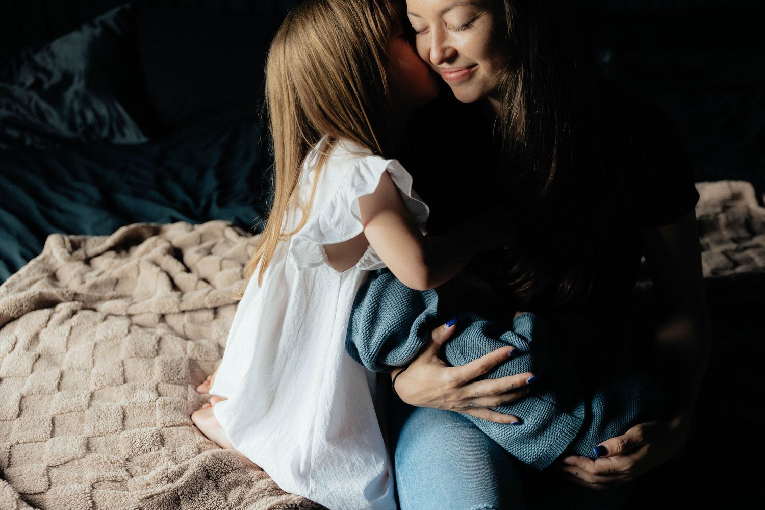 A mother and young daughter share a tender embrace while sitting on a bed with a cozy blanket in a warmly lit room.