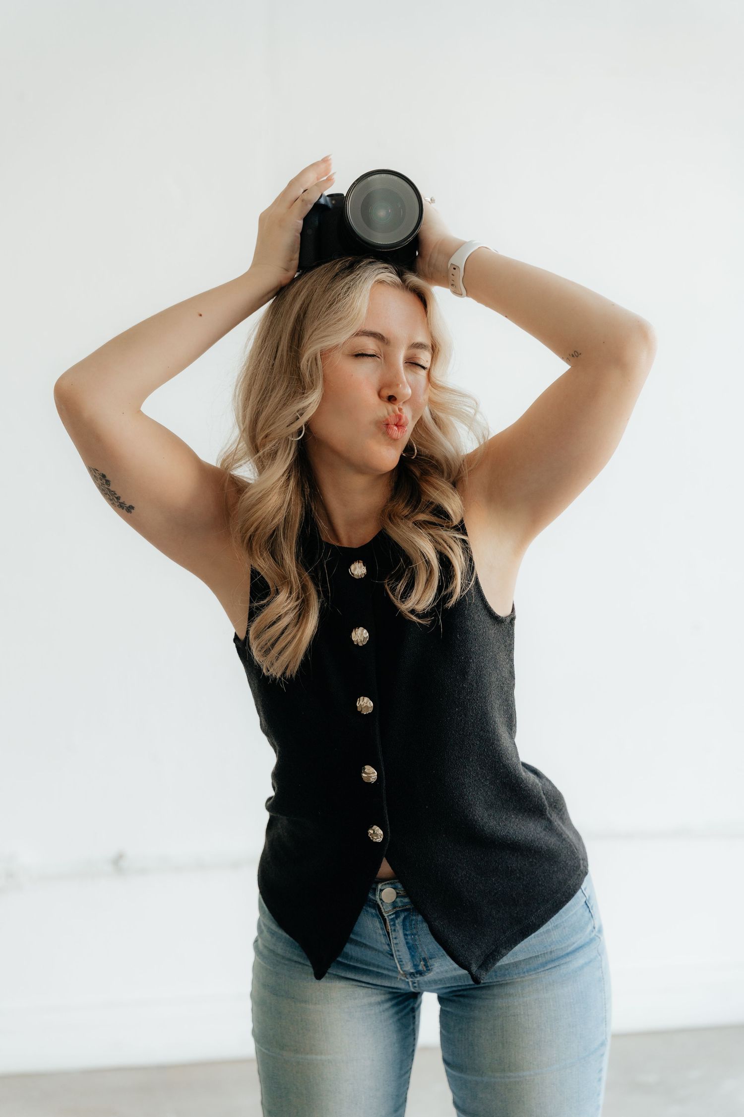 A person in a black button-up sleeveless top and jeans poses playfully against a white background.