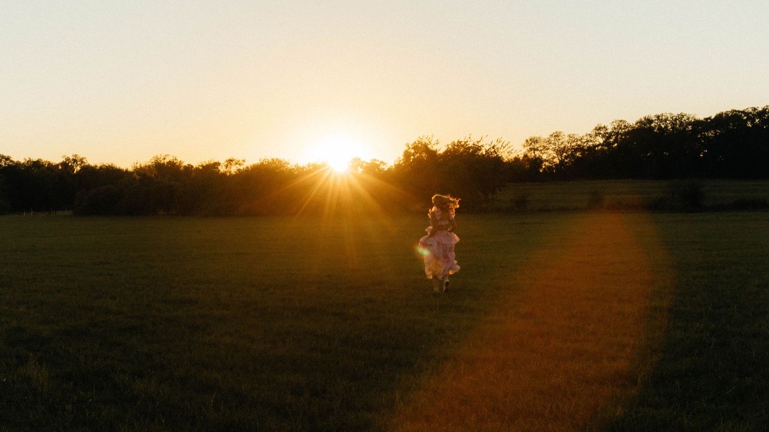 A child runs through a sunlit green field at golden hour with trees silhouetted against a glowing sunset.
