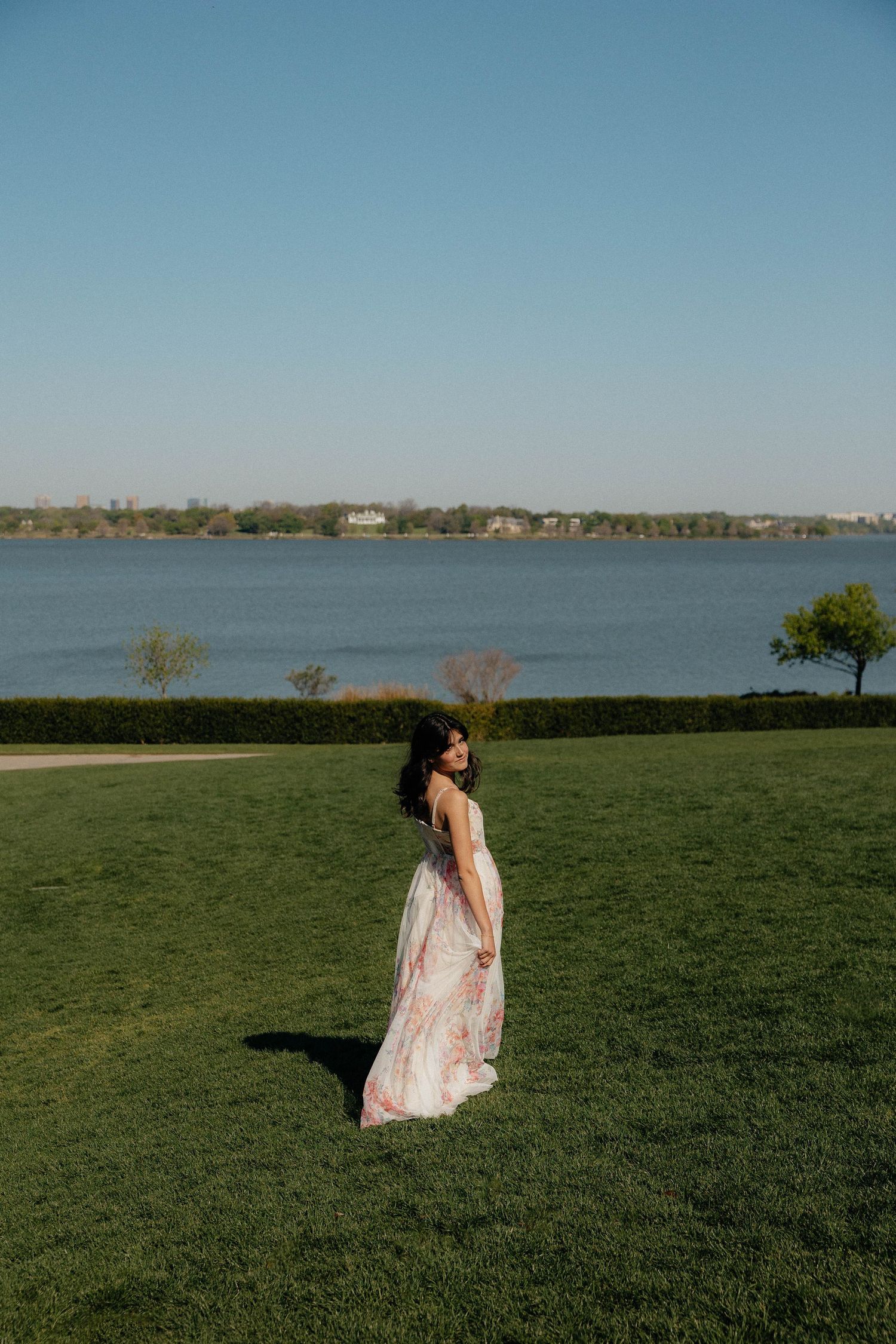 Woman in floral maxi dress standing on green lawn by waterfront on a clear sunny day.