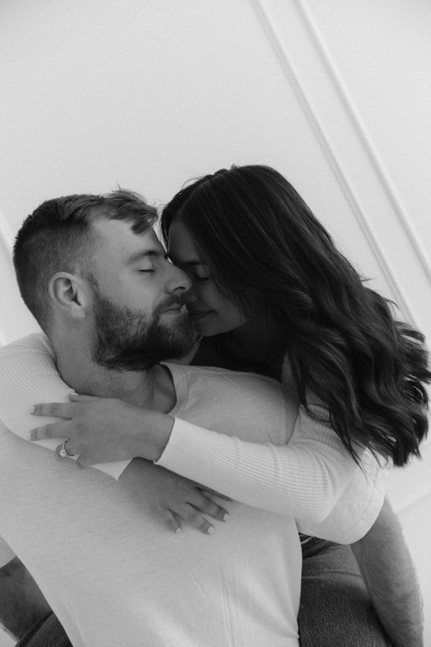 Intimate black and white portrait of a couple embracing against a white wall.
