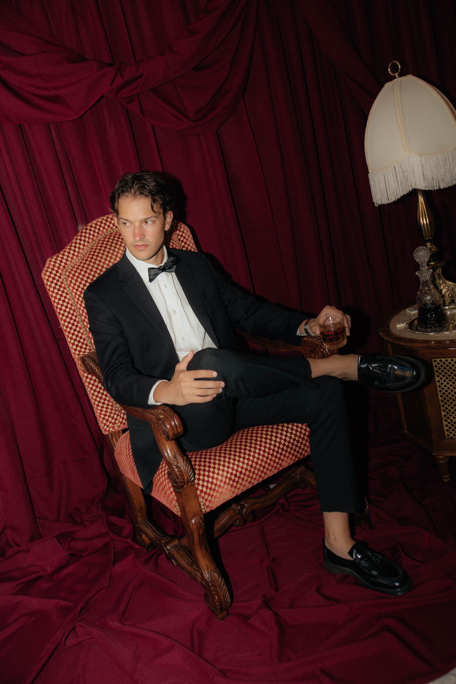 Moody portrait in formal attire against deep red velvet backdrop with vintage chair.