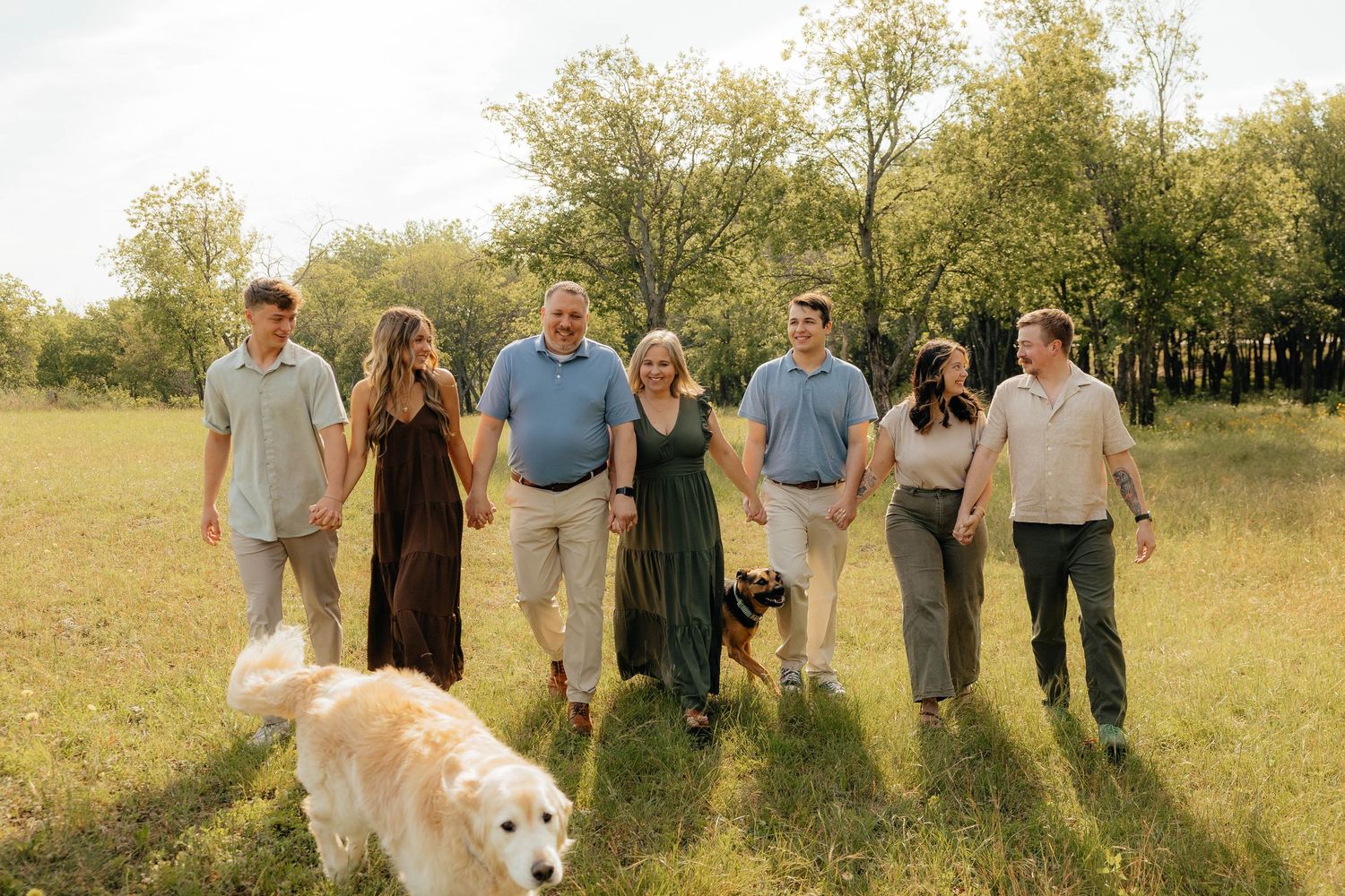 Family of seven walking through a sunny green field with two dogs, smiling and holding hands outdoors.