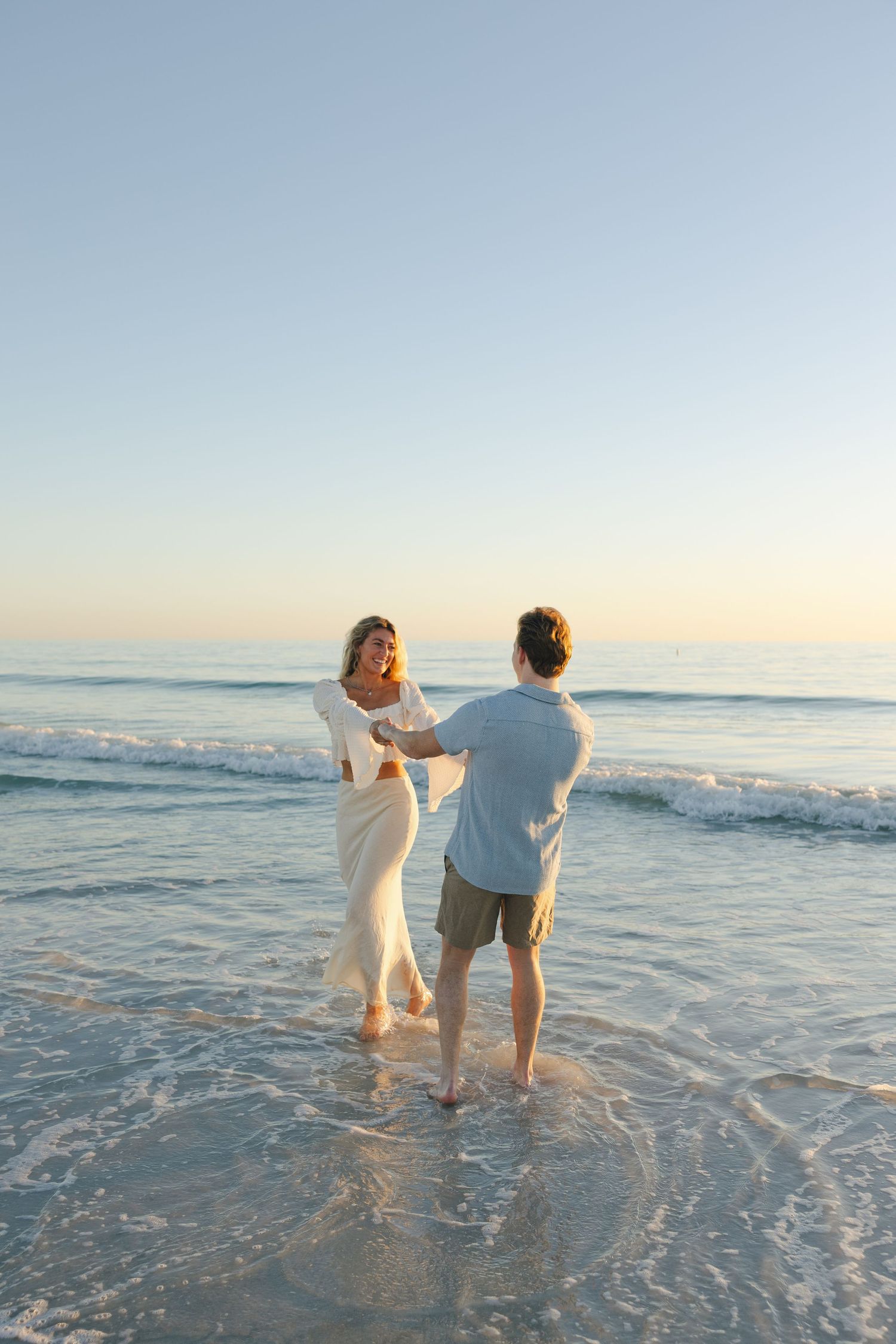 A couple stands together in shallow ocean waves at sunset on a peaceful beach.