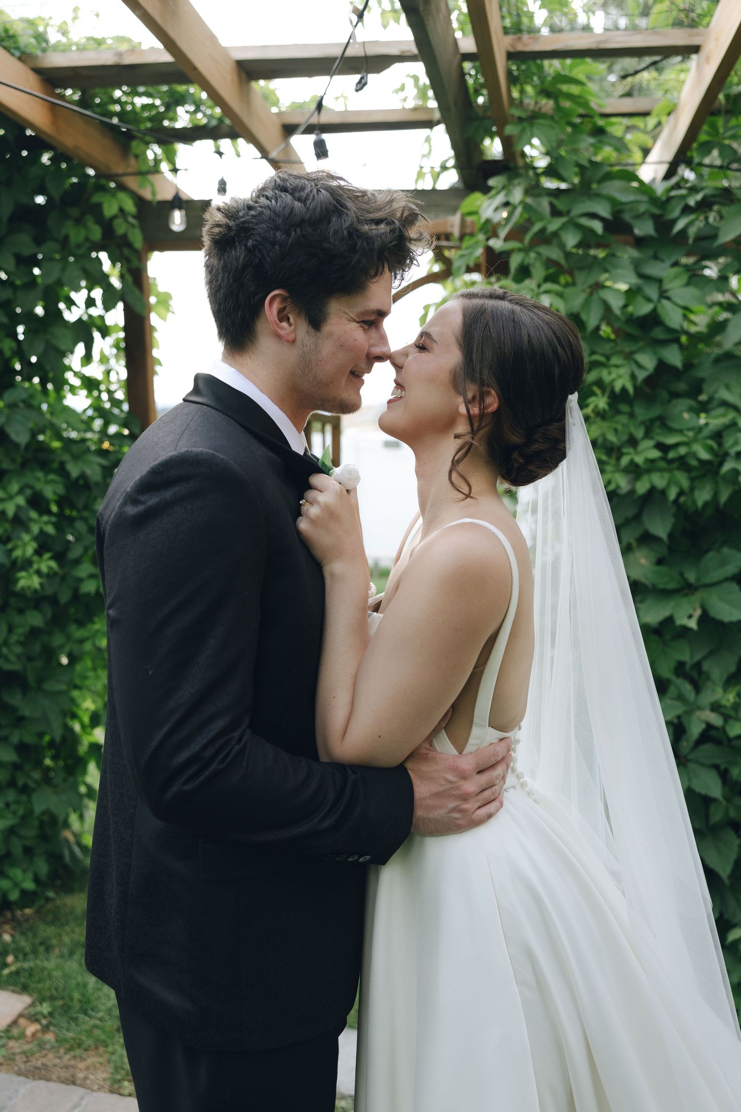 A romantic wedding couple shares an intimate kiss under a lush green garden arbor in black and white formal attire.