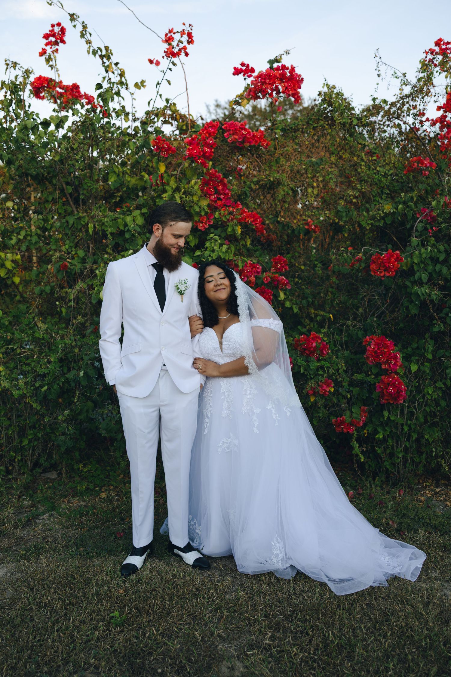 A couple in white wedding attire stands together against a backdrop of vibrant red bougainvillea flowers in bloom.