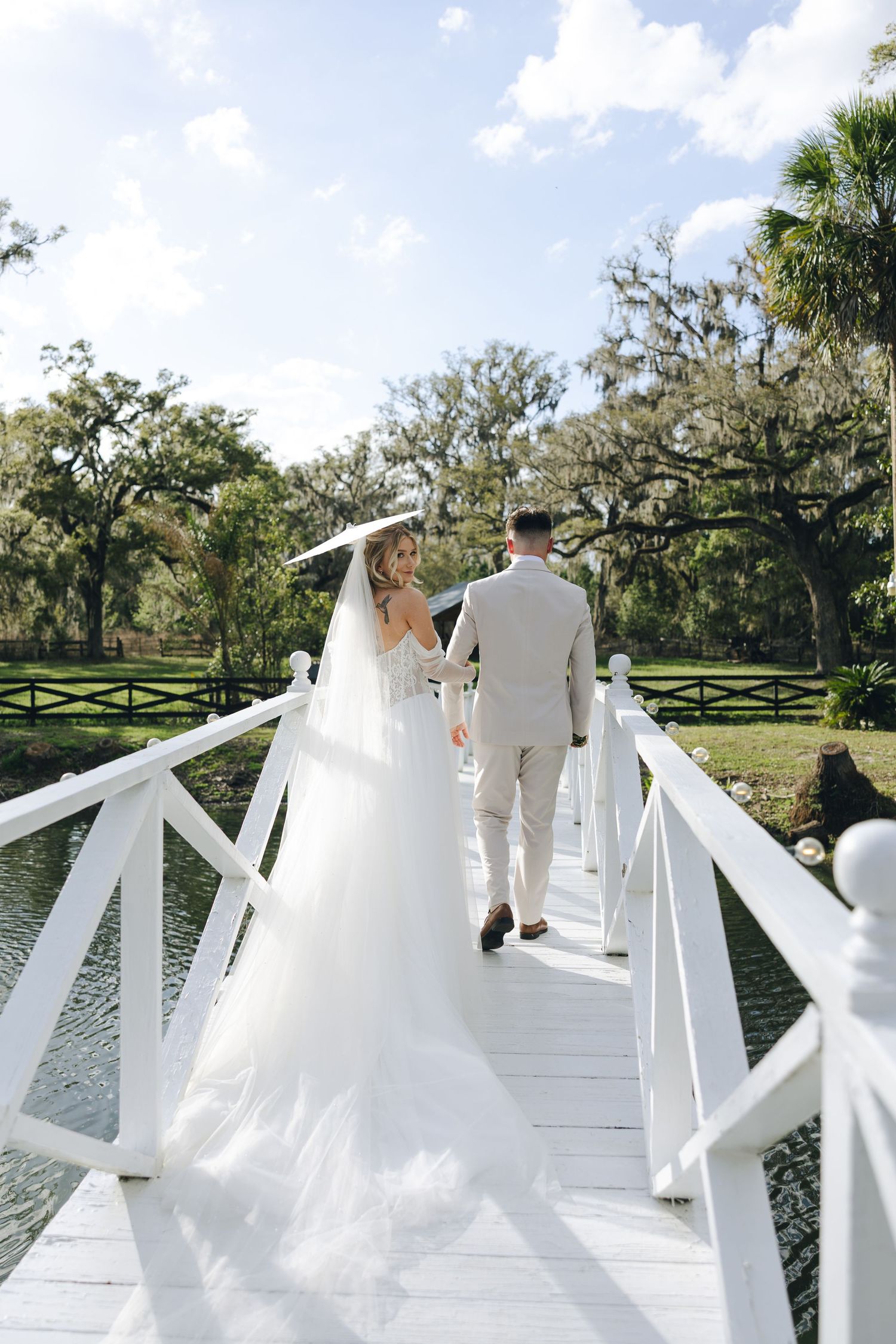 A couple in wedding attire walks down a white dock surrounded by lush greenery and water on a sunny day.