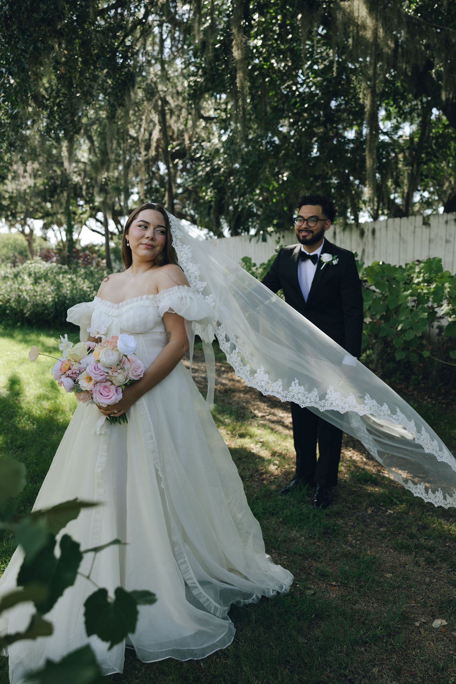 A couple in wedding attire poses outdoors in a garden setting with flowing veil and delicate flower bouquet.