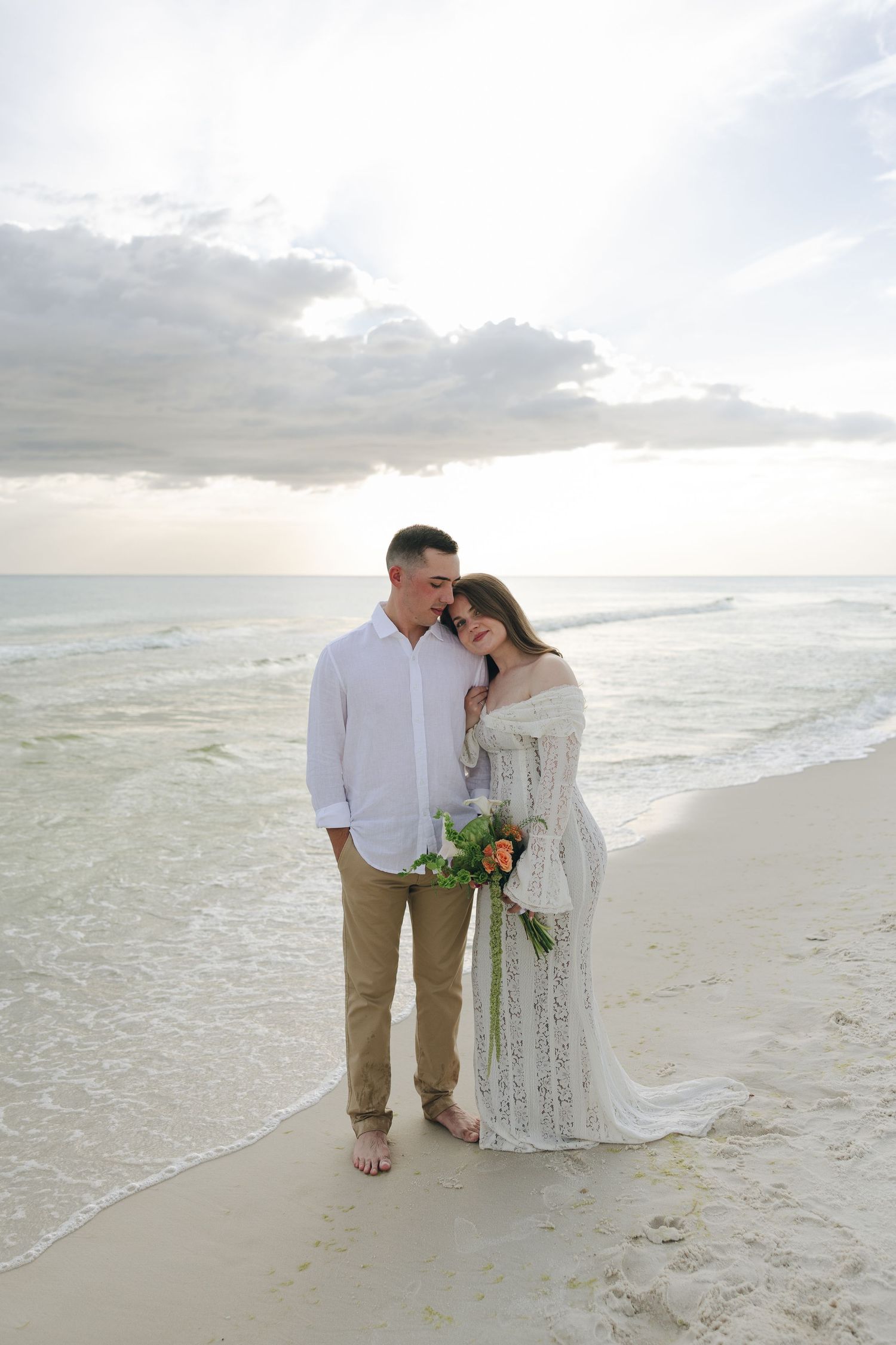 A couple in white and khaki stands together on a serene beach at sunset as waves roll in along the shoreline.