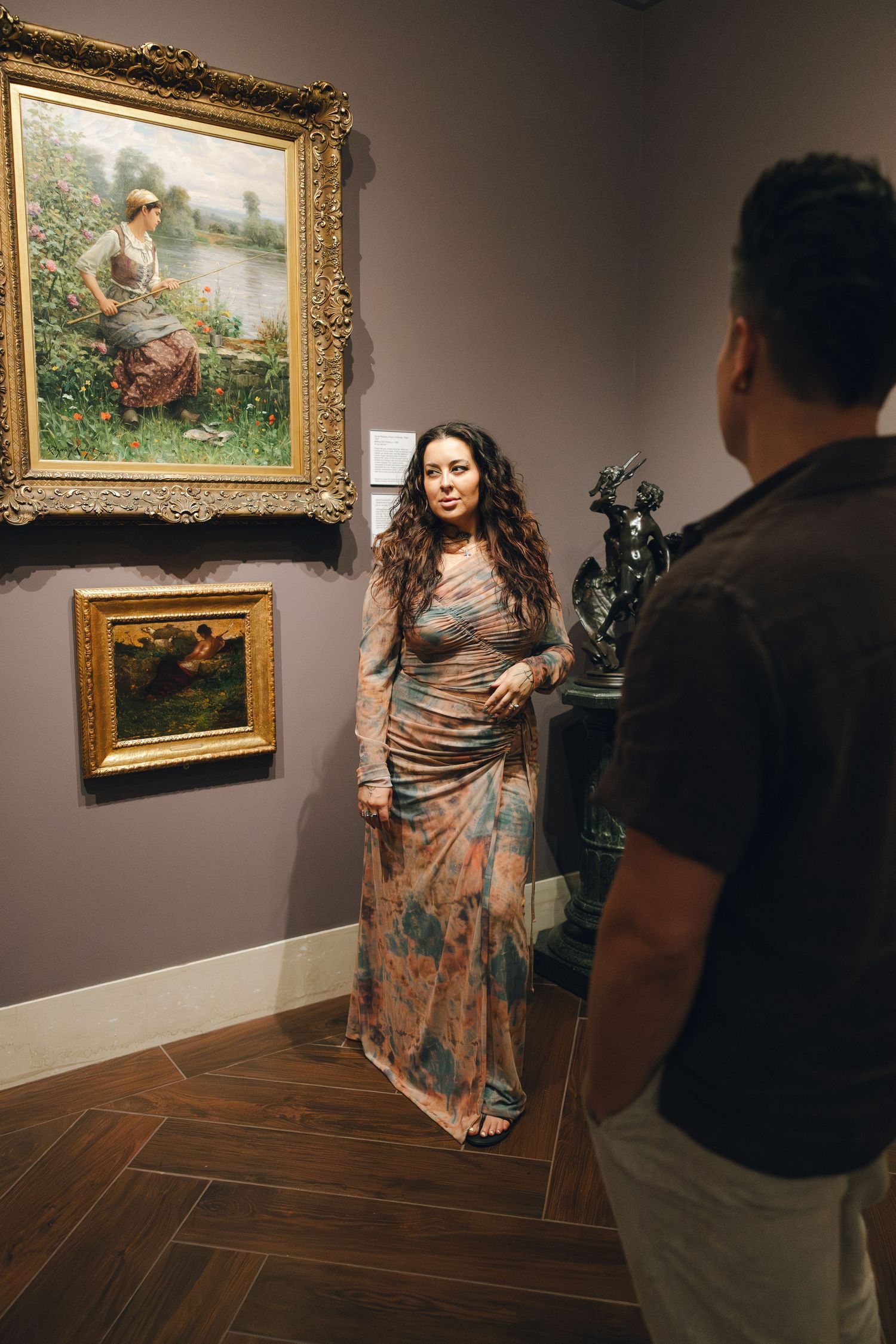 Photographer captures model posing in floral dress against gallery wall displaying classic paintings in museum setting.