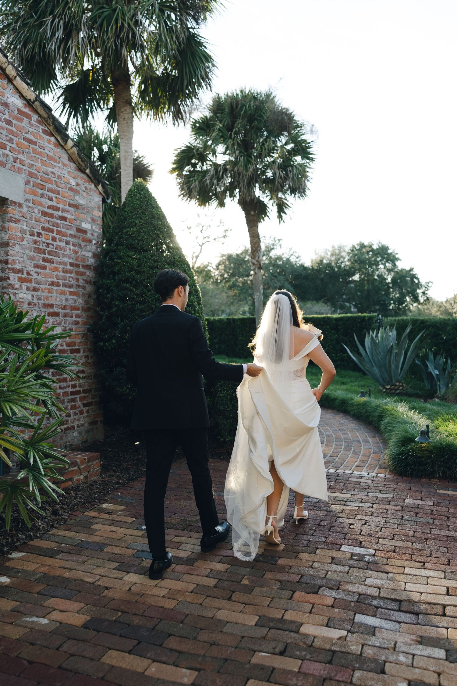 A couple in wedding attire walks along a brick path surrounded by palm trees and tropical landscaping.