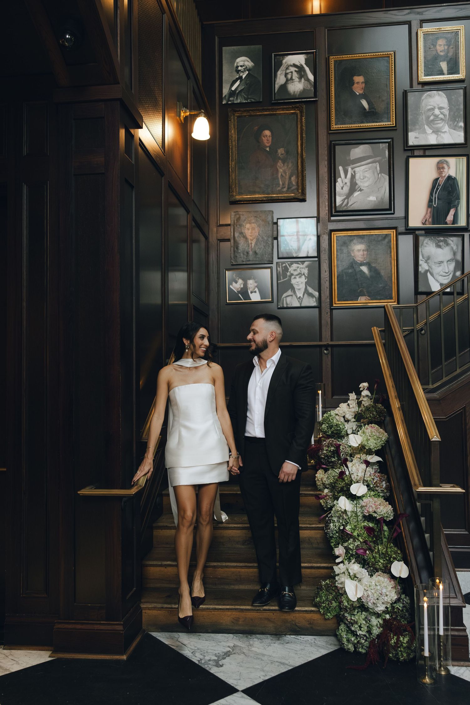 Stylish couple stands on elegant staircase with vintage portrait gallery wall and floral arrangements in moody lighting.