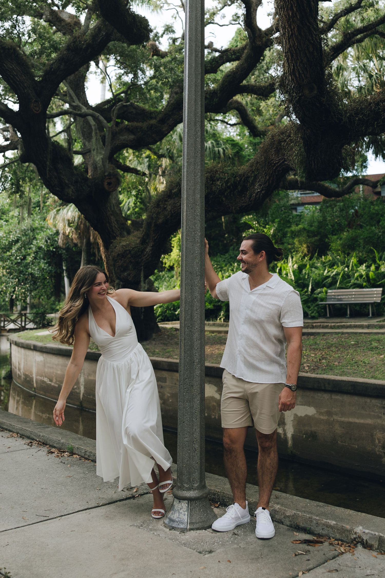 A playful lifestyle photoshoot of a couple in white outfits posing along a park pathway with oak trees in the background.