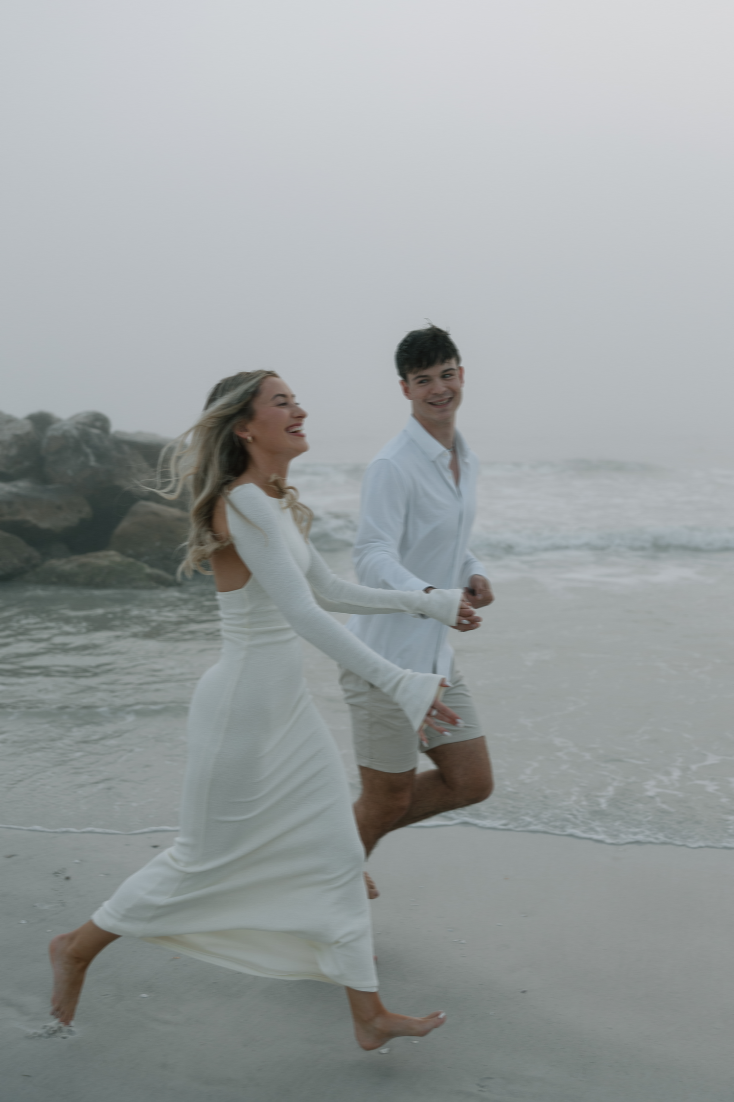 A joyful couple runs along a misty beach in white clothing.
