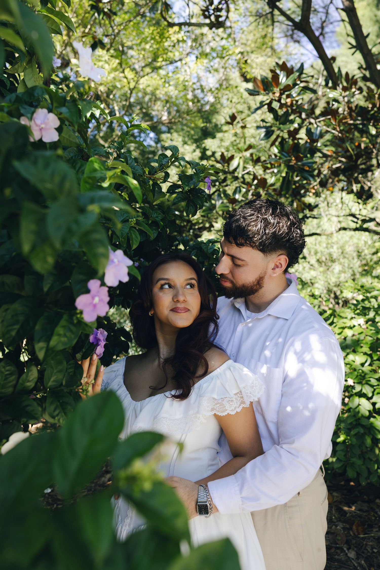A romantic couple in white outfits embrace among blooming flowers and greenery in a garden setting.