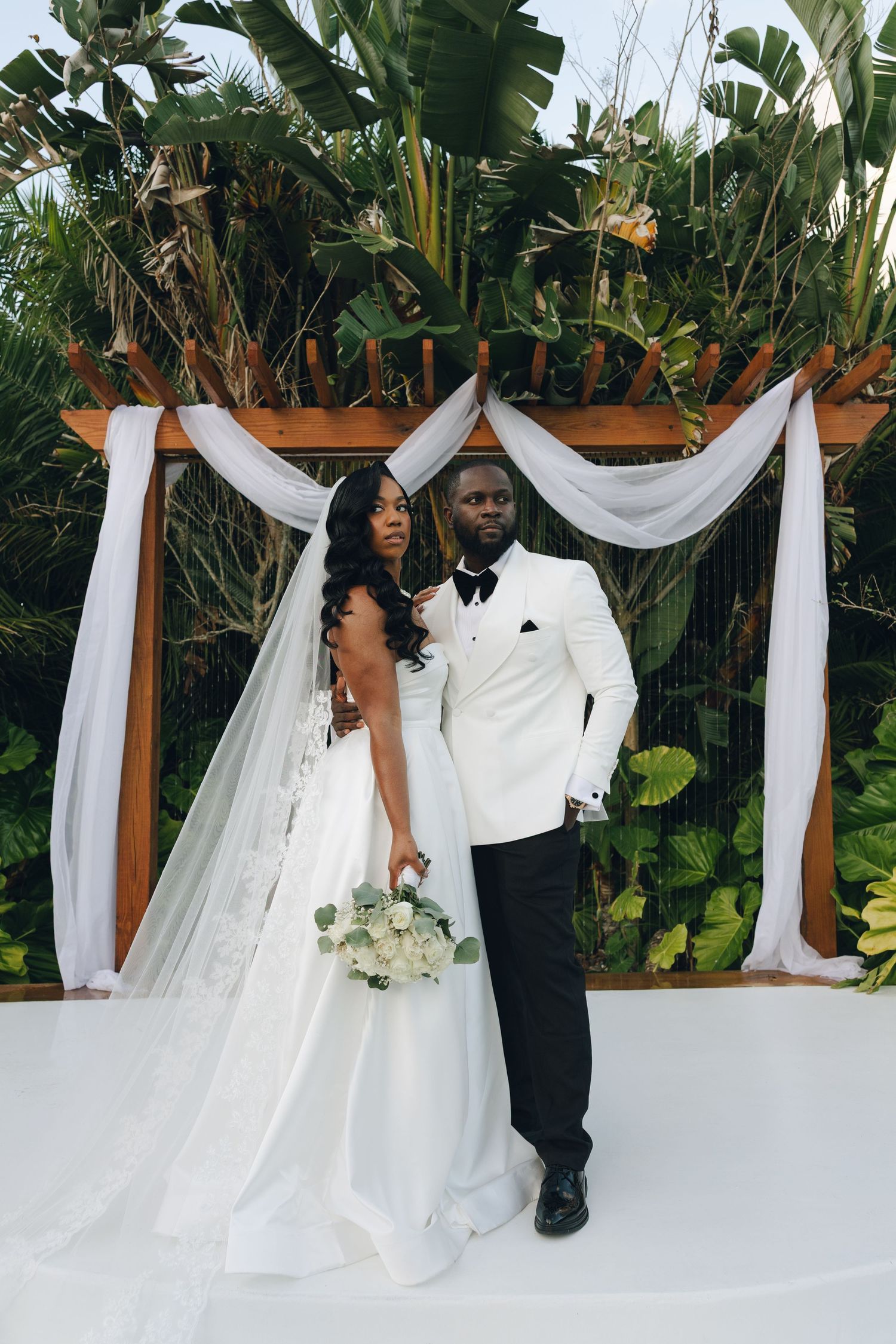 A couple shares a joyful moment during their wedding ceremony under a white draped arch with tropical foliage backdrop.